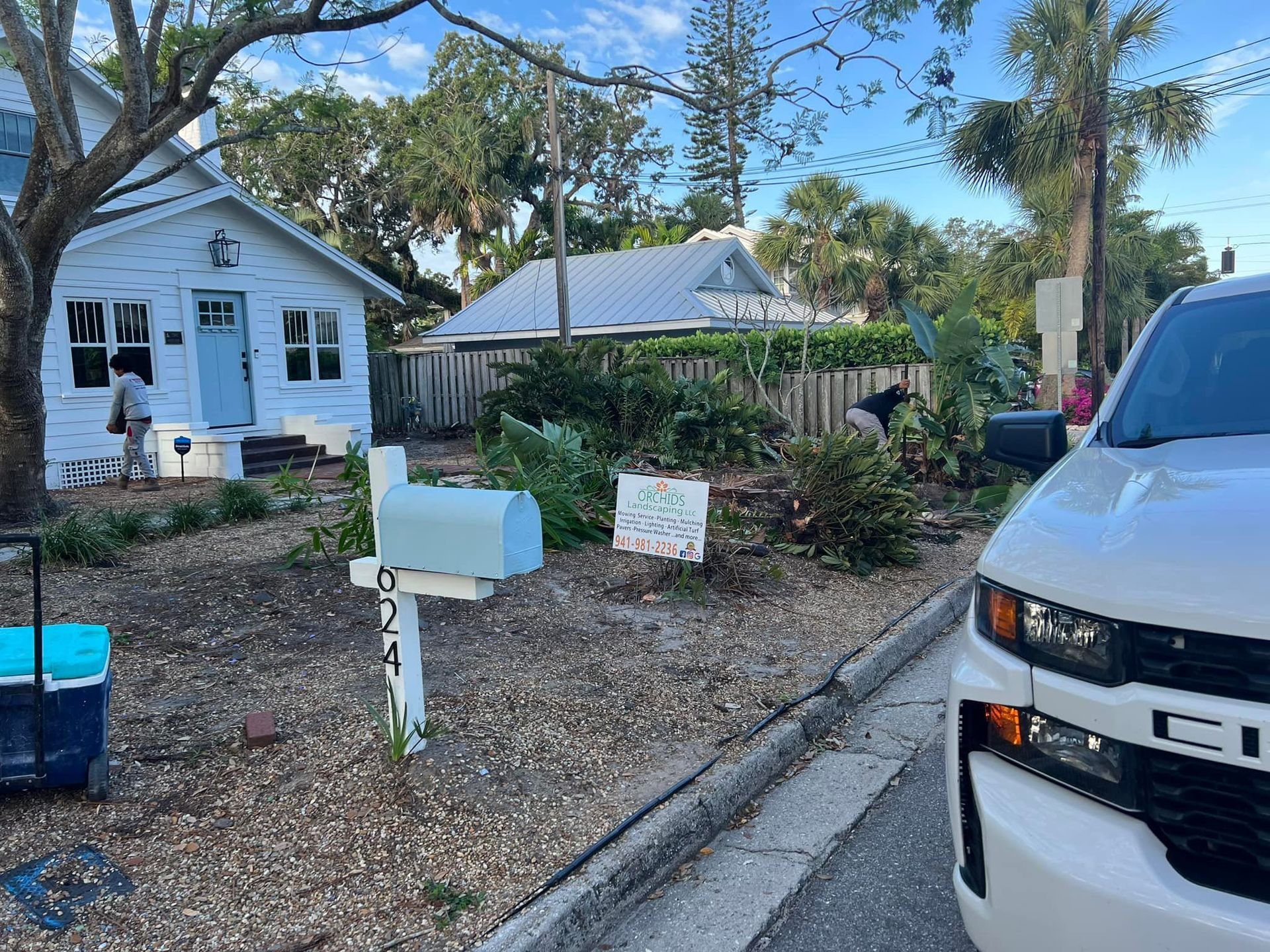Blue mailbox in front yard with small house, white car, and person gardening.