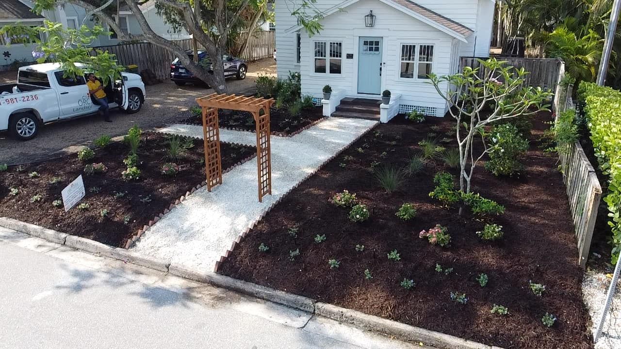 White house with blue door, landscaped yard, gravel path leading to an arch, and a truck parked to the side.
