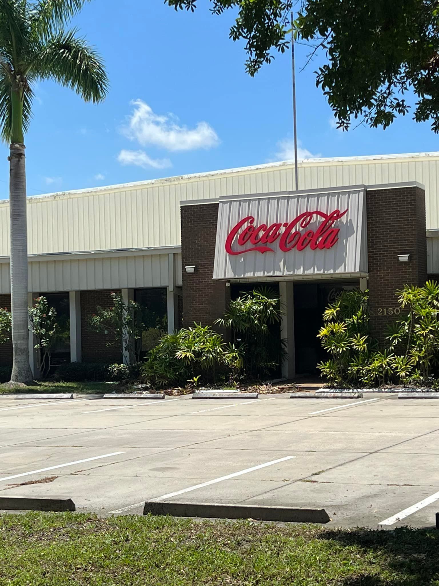 Coca-Cola building with red logo, brick facade, and a parking lot on a sunny day.