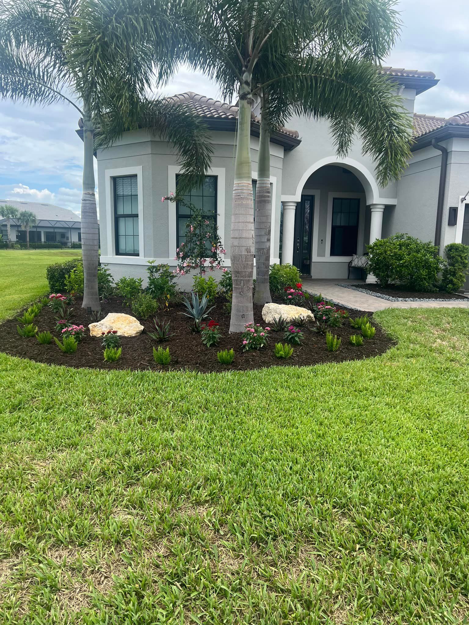 A house with a manicured lawn and garden. Palm trees frame the entryway, beds of flowers and rocks are in front.