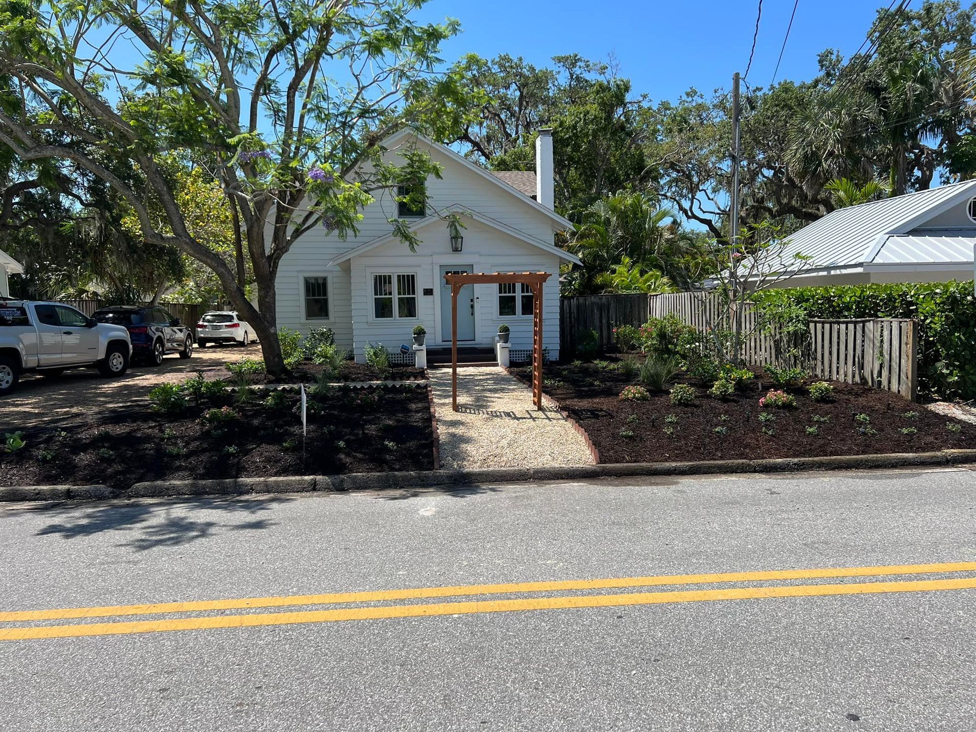 White house with stone path, arbor, and landscaping. Cars parked on the left; street in foreground.