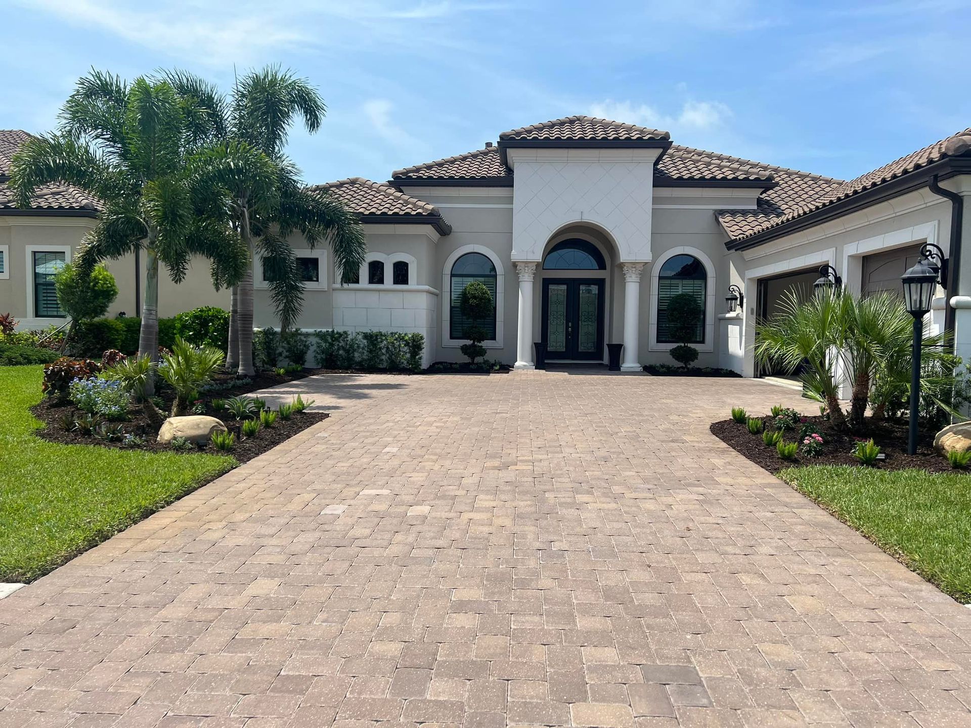 Beige brick paved driveway leading to a large light-colored house with a tiled roof and manicured landscaping under a blue sky.