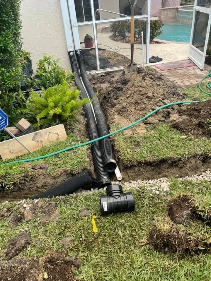 A drainage system under construction near a house and pool. Black corrugated pipes, dirt, and tools are visible.