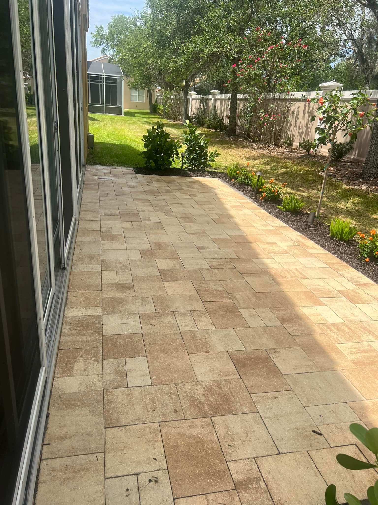 Patio with beige pavers, plants along the edge, and a glimpse of a house and trees in the background.