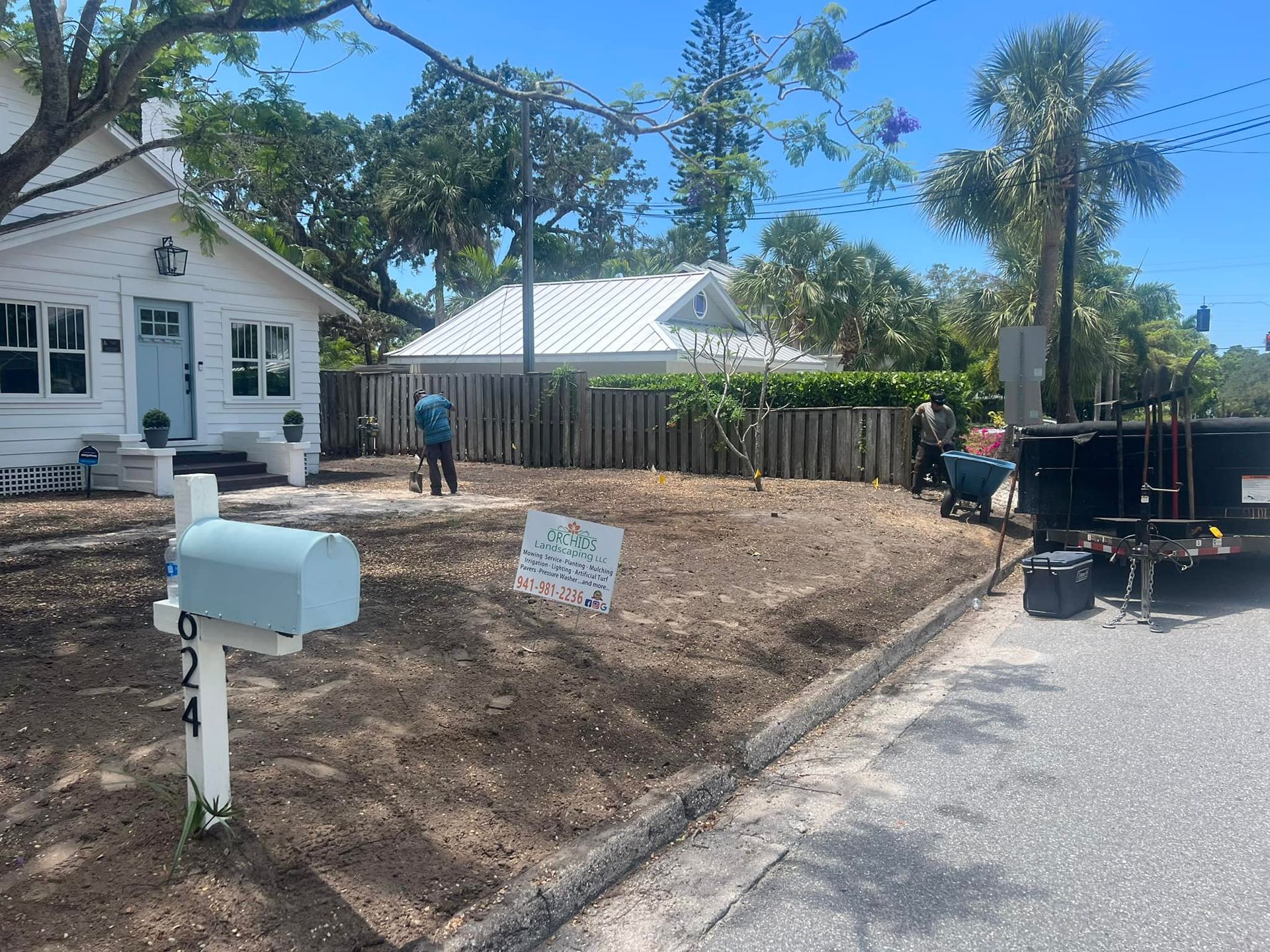 Yard work in progress at a white house; people working on landscaping. A blue mailbox stands in the foreground.