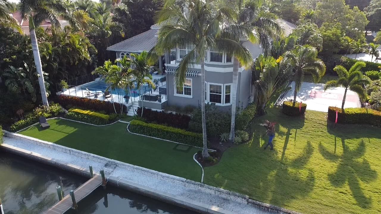 Luxury house with pool, dock, and palm trees. Gray exterior, green lawn, sunny day.