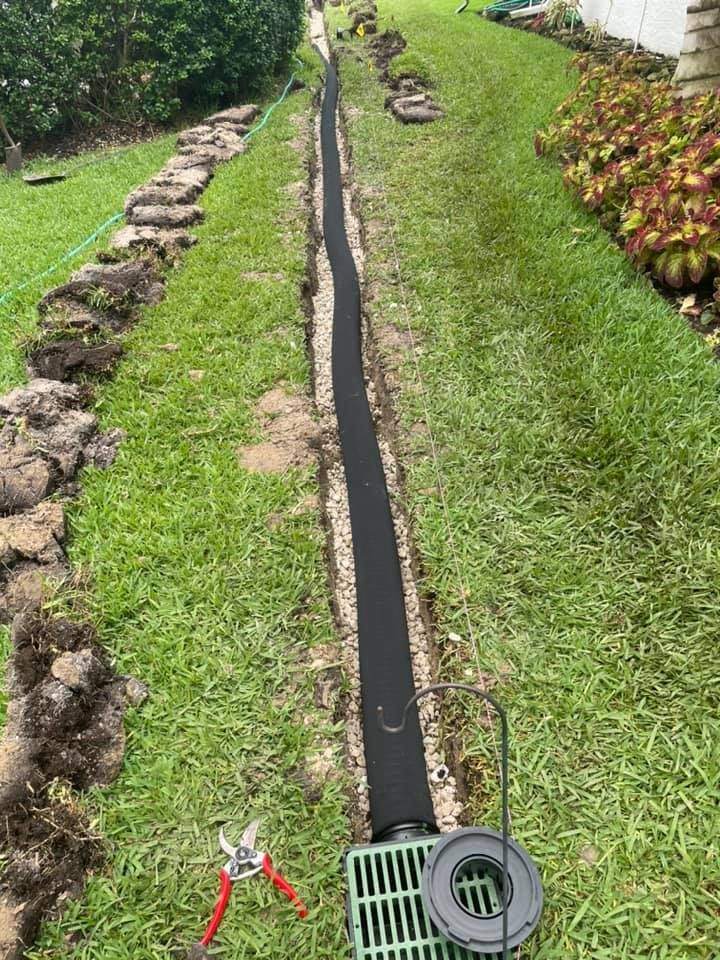 A long, narrow trench with black fabric and gravel. A green grate and coiled wire are at the bottom.