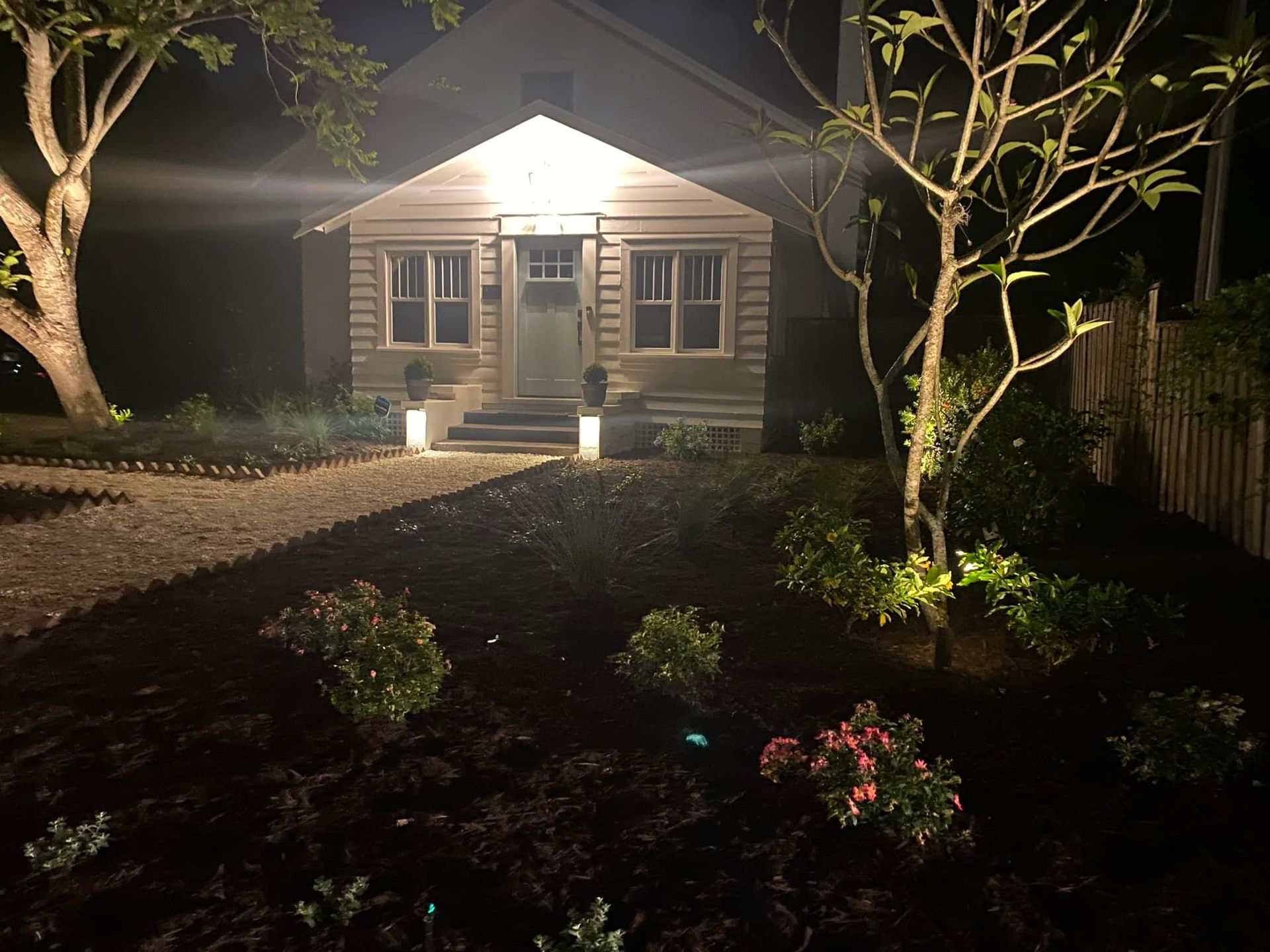 A small house at night, lit by spotlights on front yard landscaping.