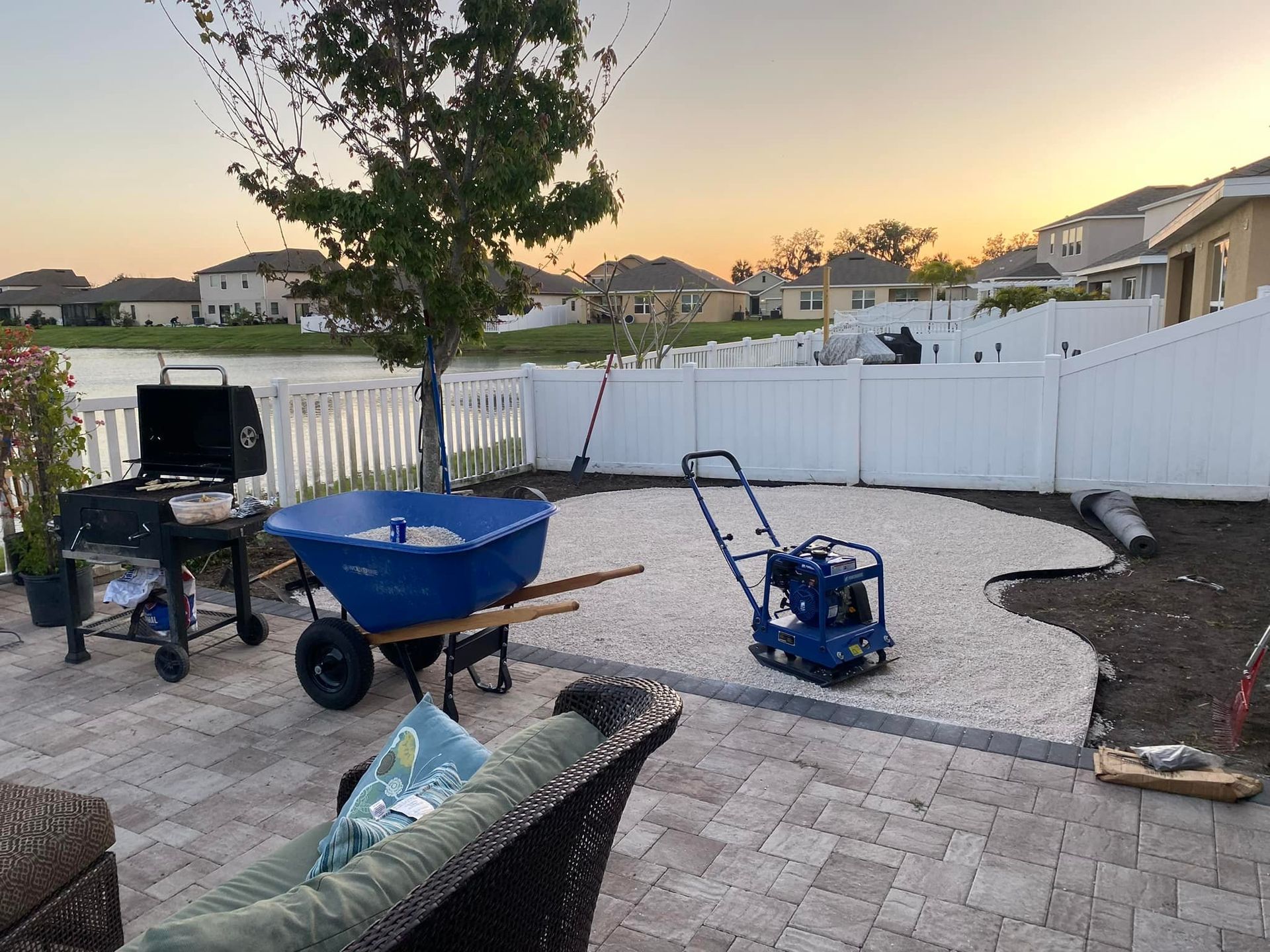Backyard scene: a gravel area under construction, with wheelbarrow, compactor, grill, and fence. Lake in the background.