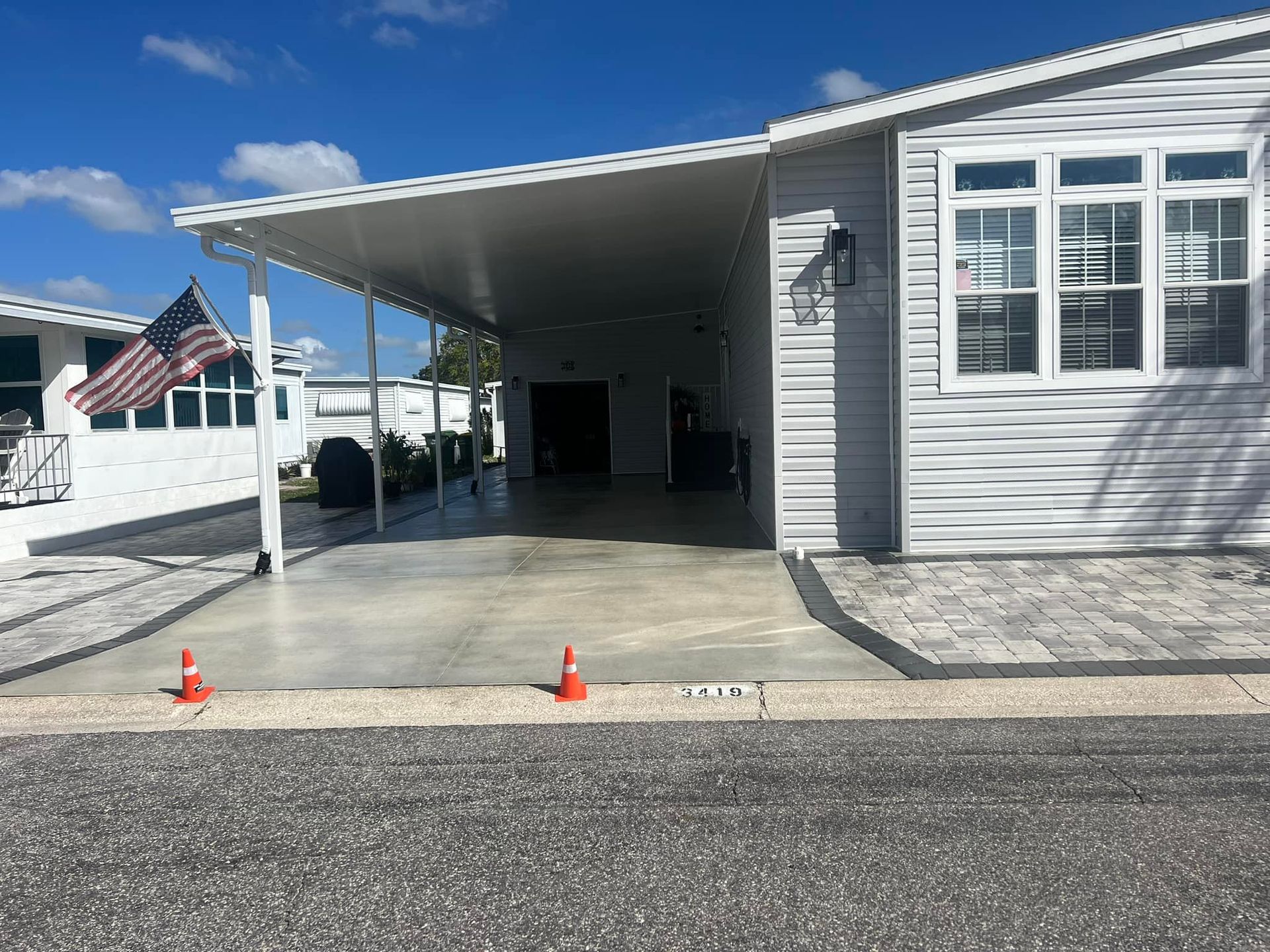 Carport of a light gray mobile home with a white awning and an American flag.