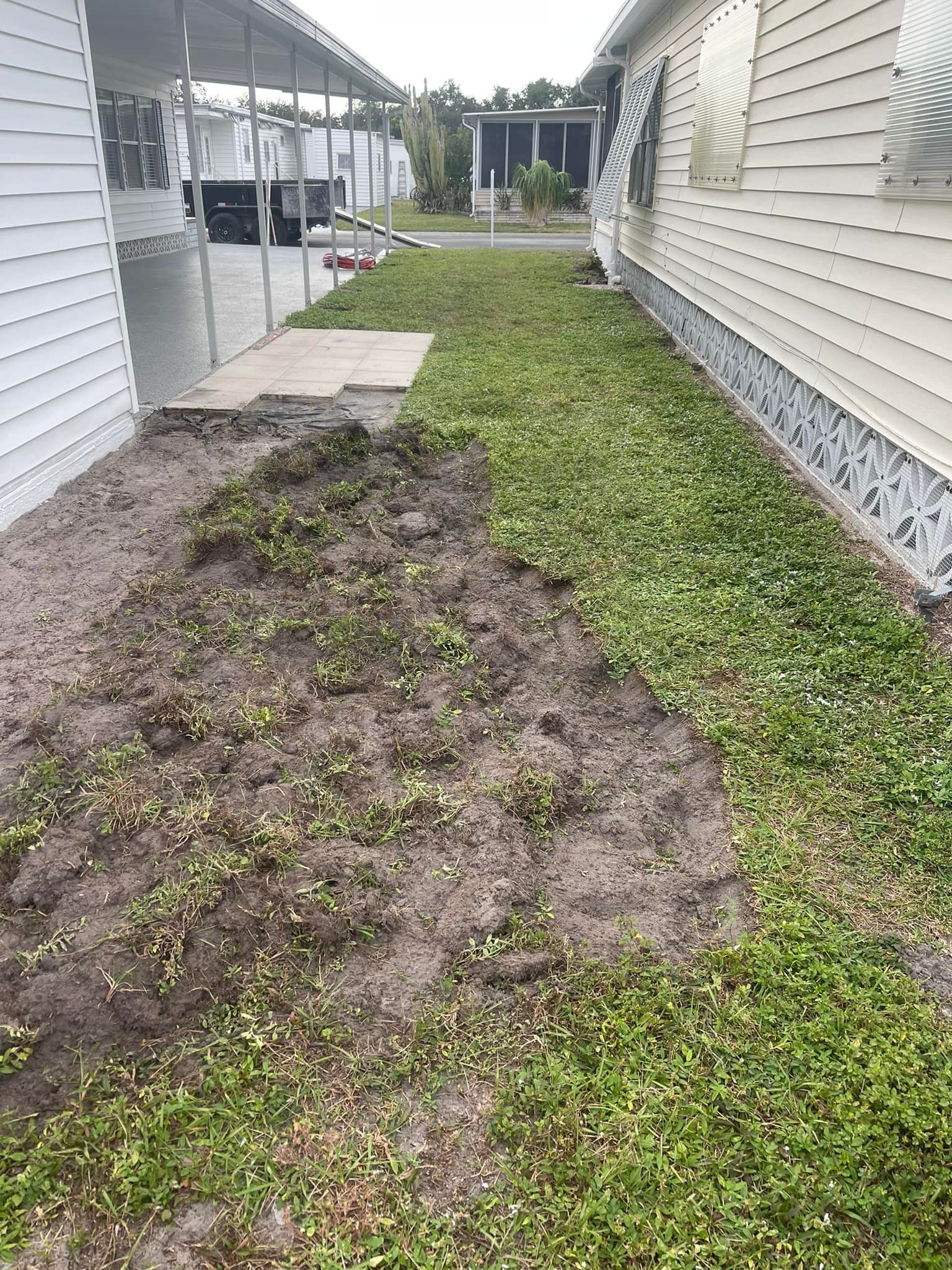 Dirt patch next to a building, with overgrown grass. Gray siding, overcast sky.