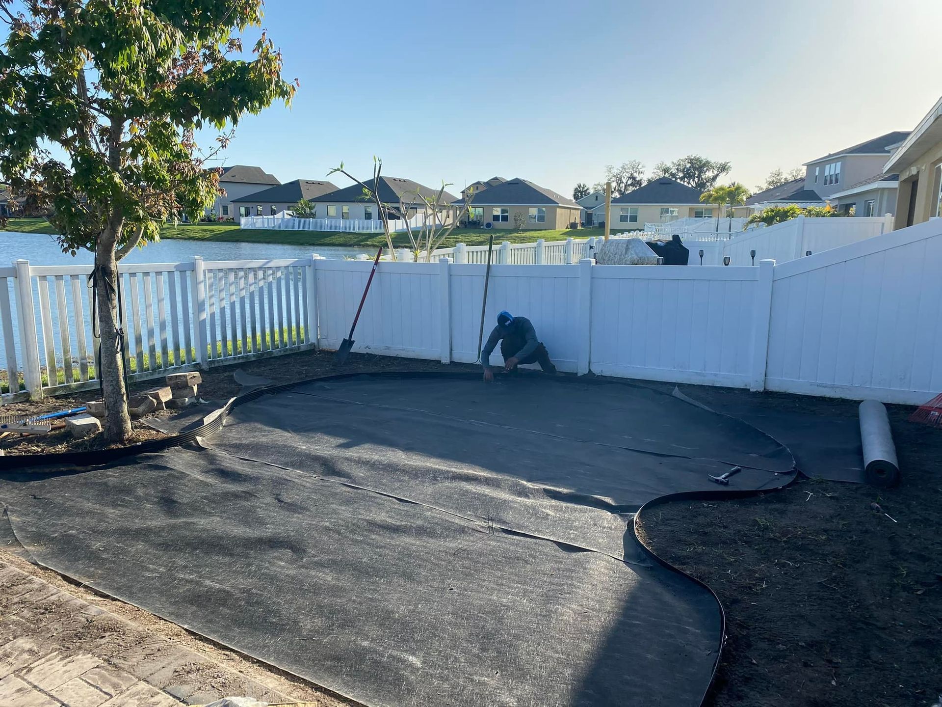 Backyard landscaping with fence, lake, mulch, and worker installing fabric.