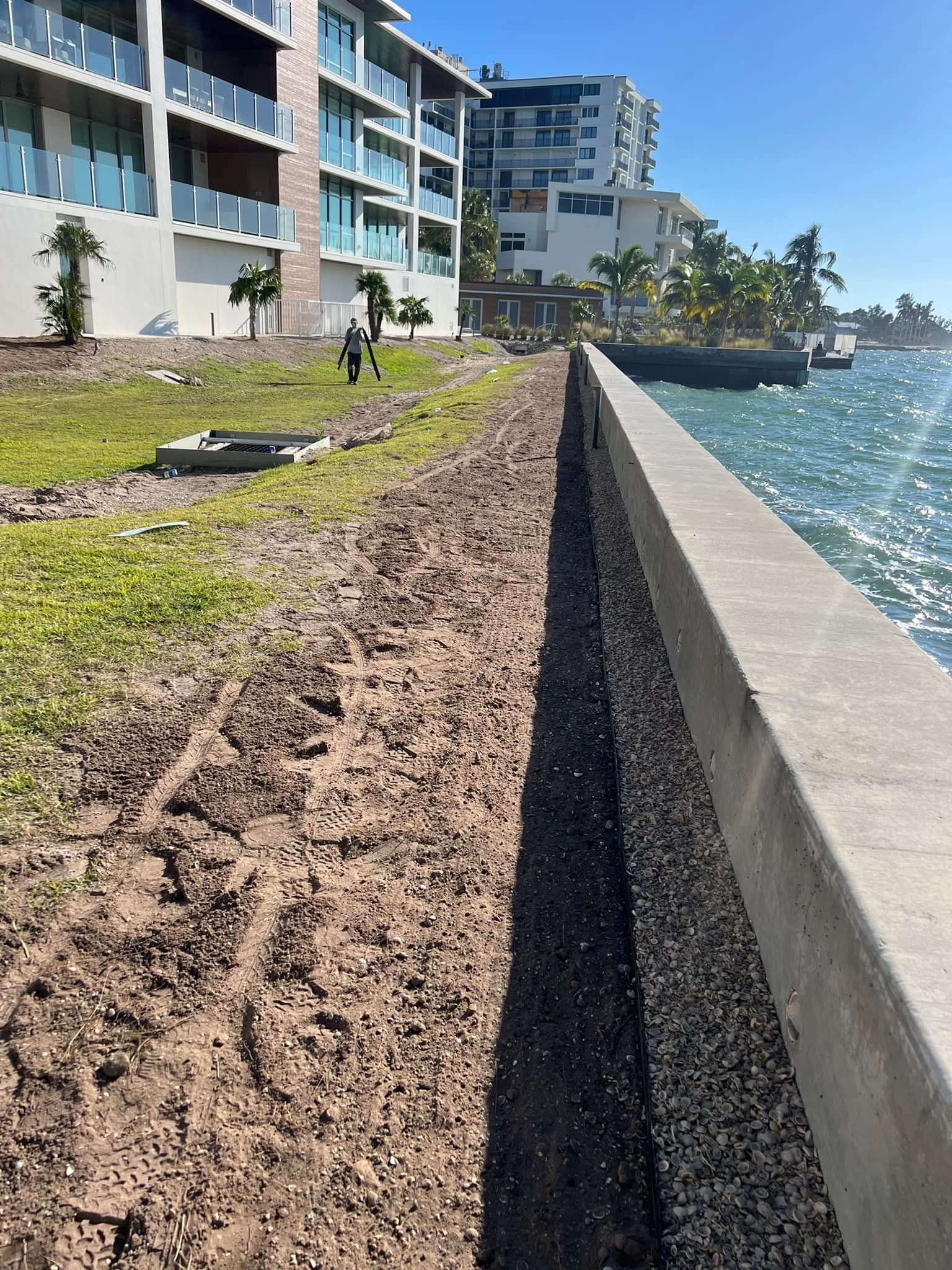 Concrete seawall next to water. Brown earth and grass on left. Buildings in background.