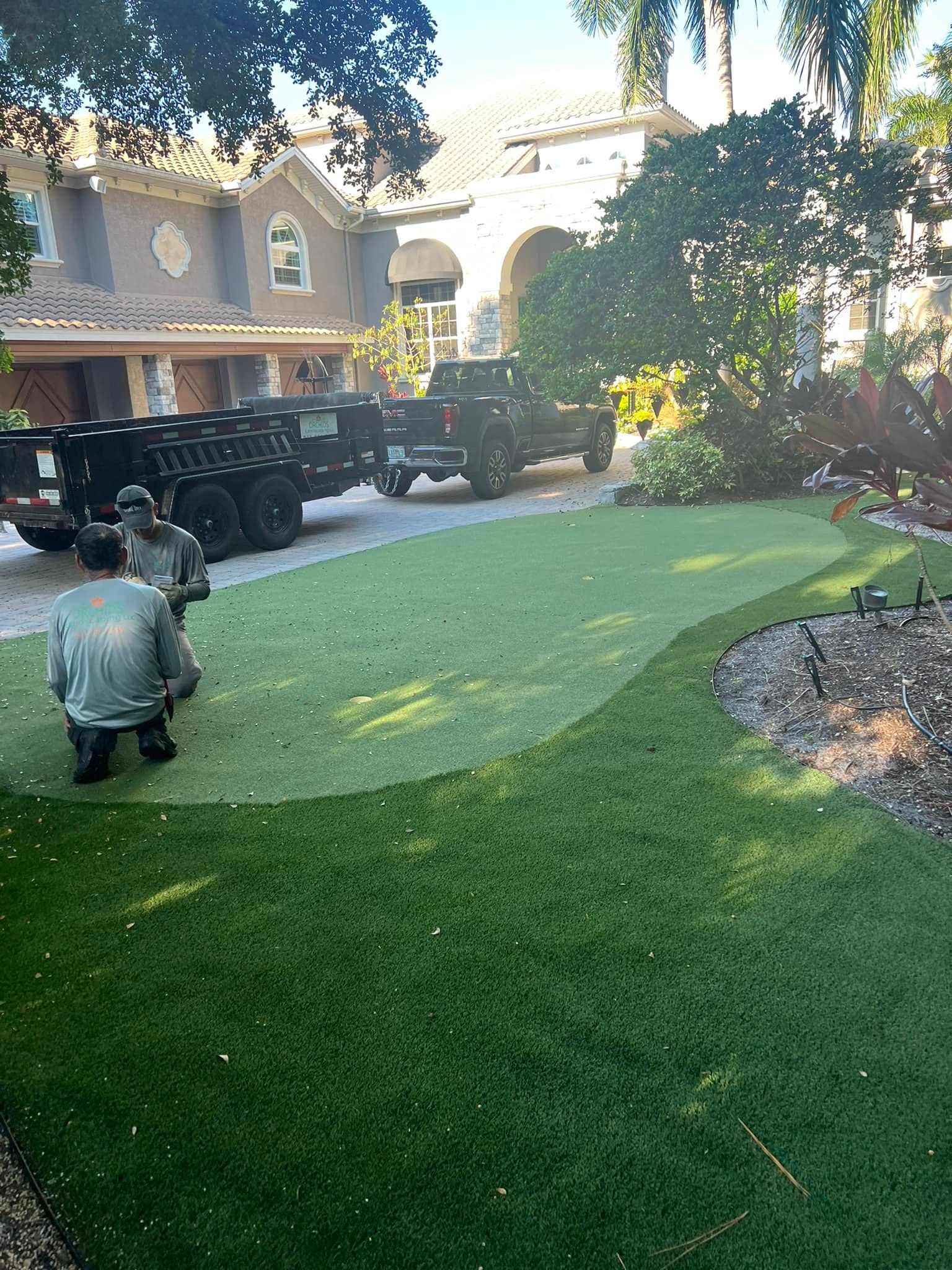 Men installing artificial turf in front of a large house, with a truck and trailer parked nearby.