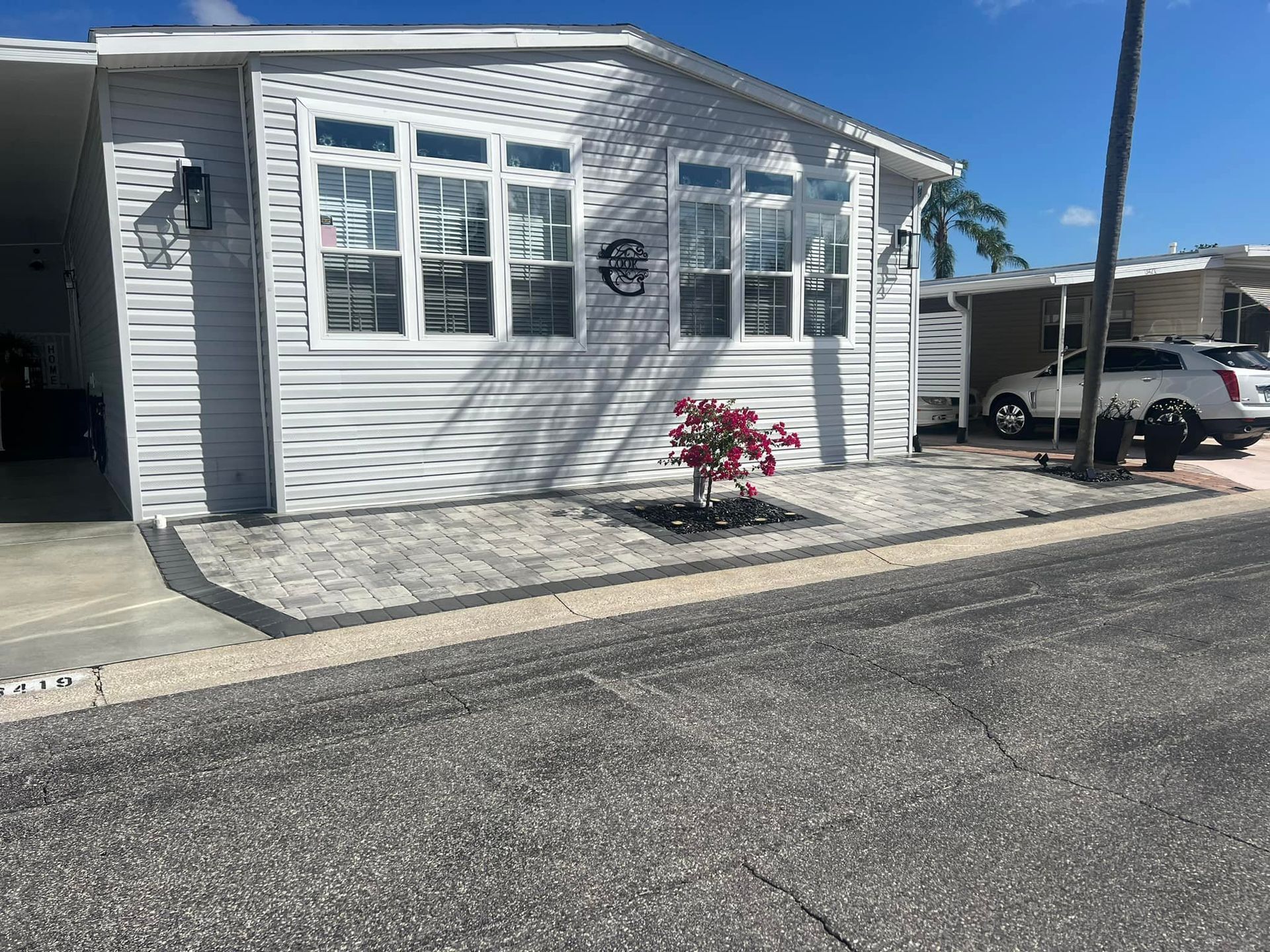 Gray manufactured home with white-framed windows, red flowers, and a car parked nearby on a sunny day.