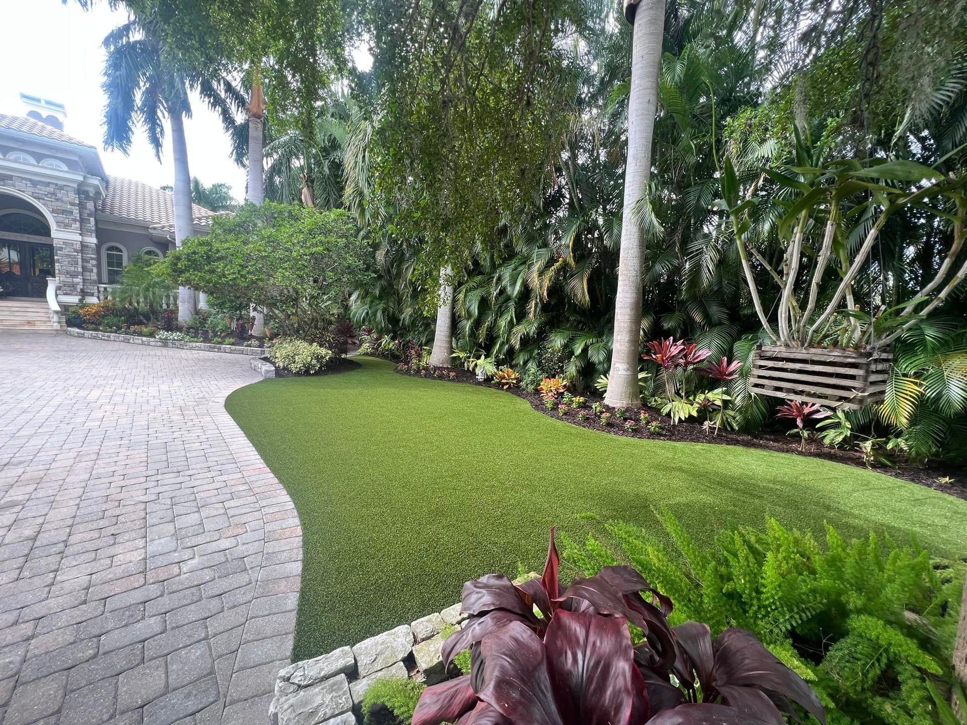 Brick driveway curves past manicured lawn and lush tropical landscaping.
