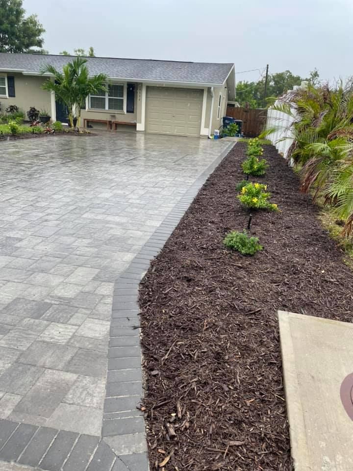Driveway with paver design, bordering a mulch garden bed with small plants in front of a house.