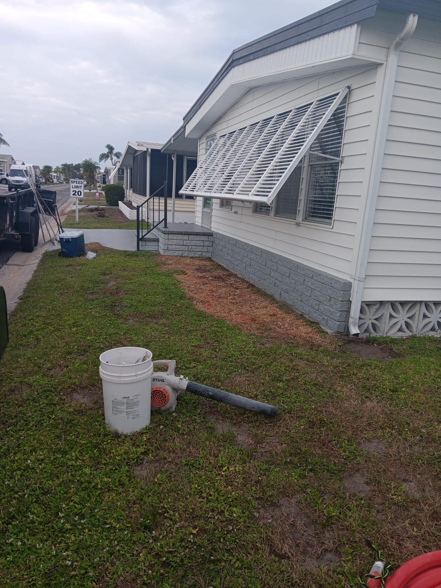 A white house with awning, a bucket, and a leaf blower on the grass.