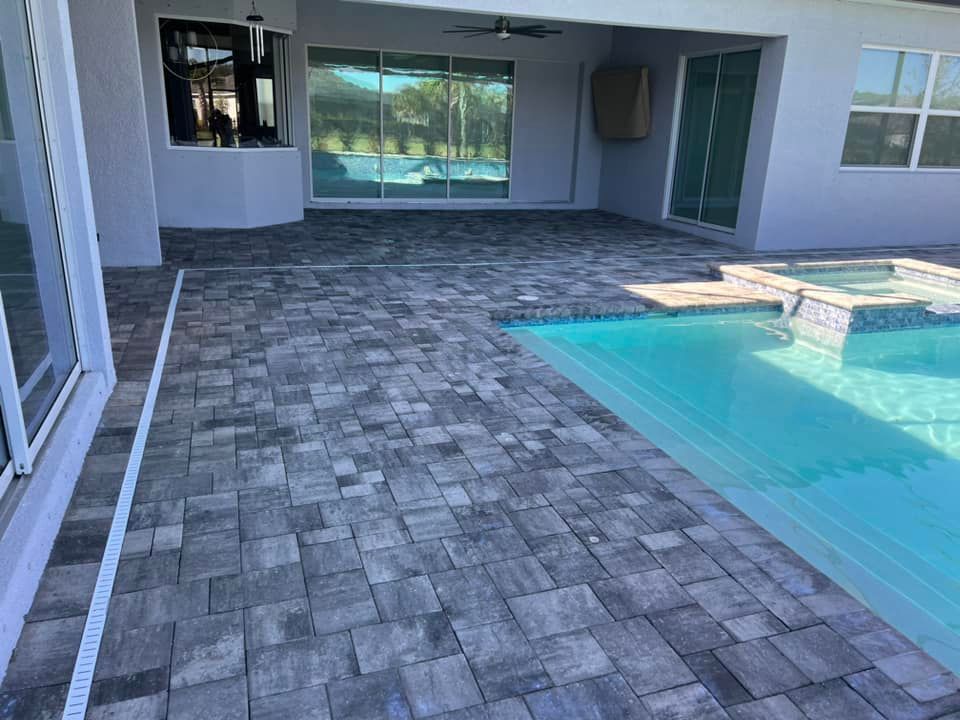 Poolside patio with gray pavers and a turquoise pool. A covered area with sliding glass doors is visible.