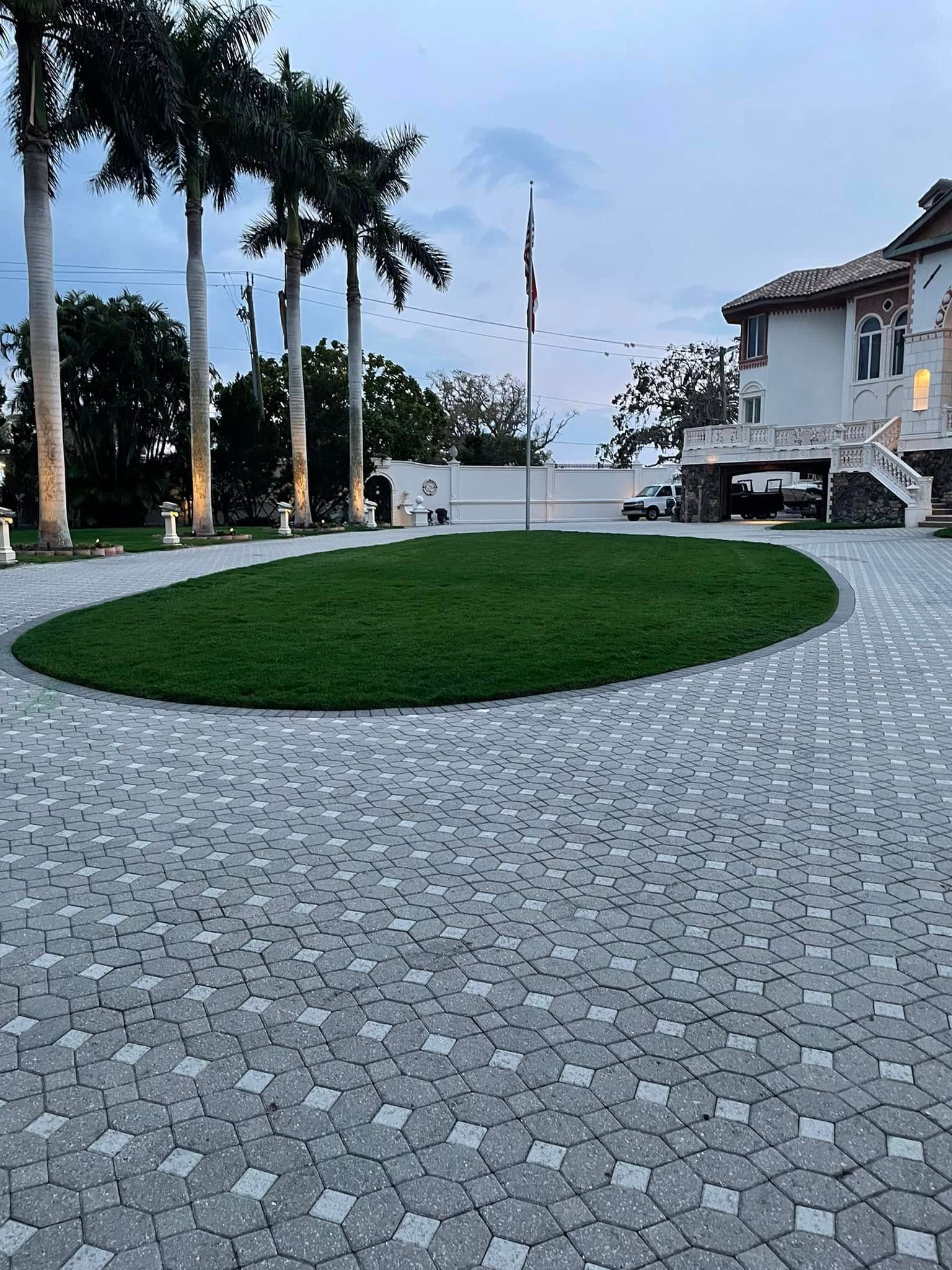 Brick driveway curves around a green grassy patch with palm trees and a white building in the background.