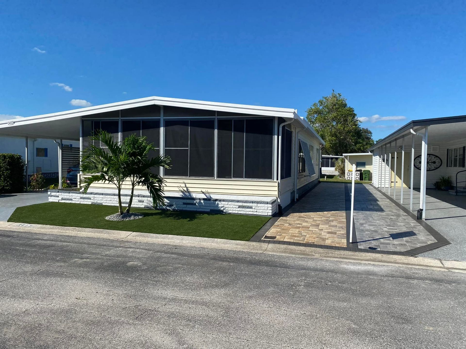 Mobile home with white exterior, screen porch, artificial turf lawn, and patterned walkway on a sunny day.