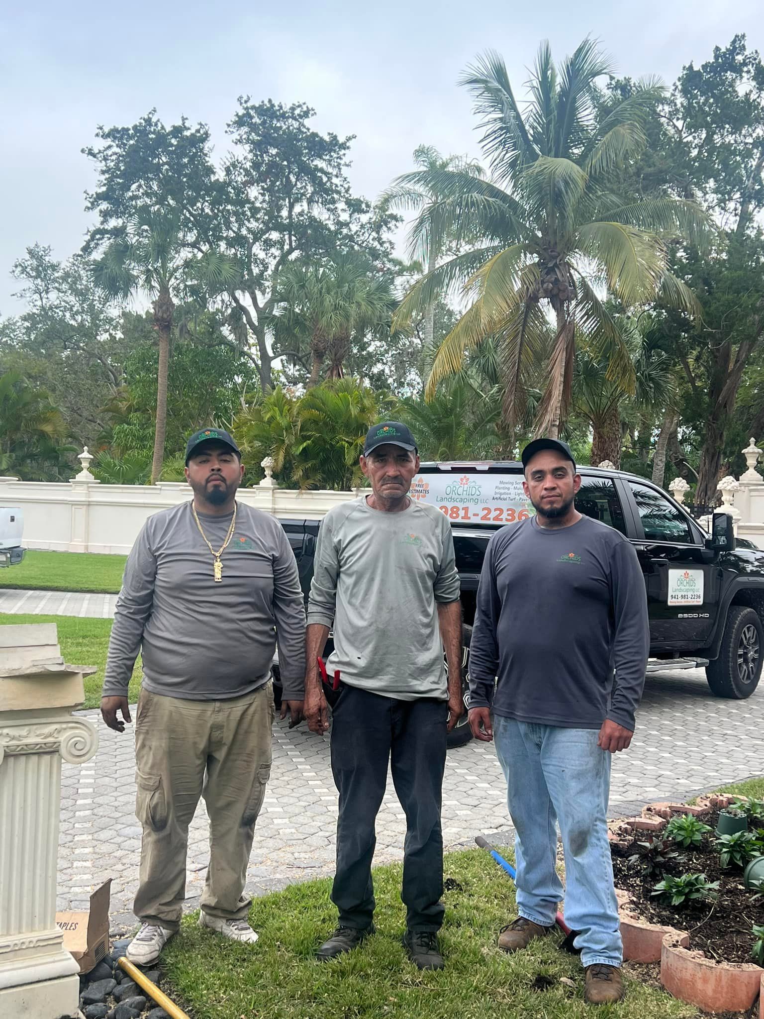 Three men in work clothes stand in front of a truck and landscaping, possibly a construction site.