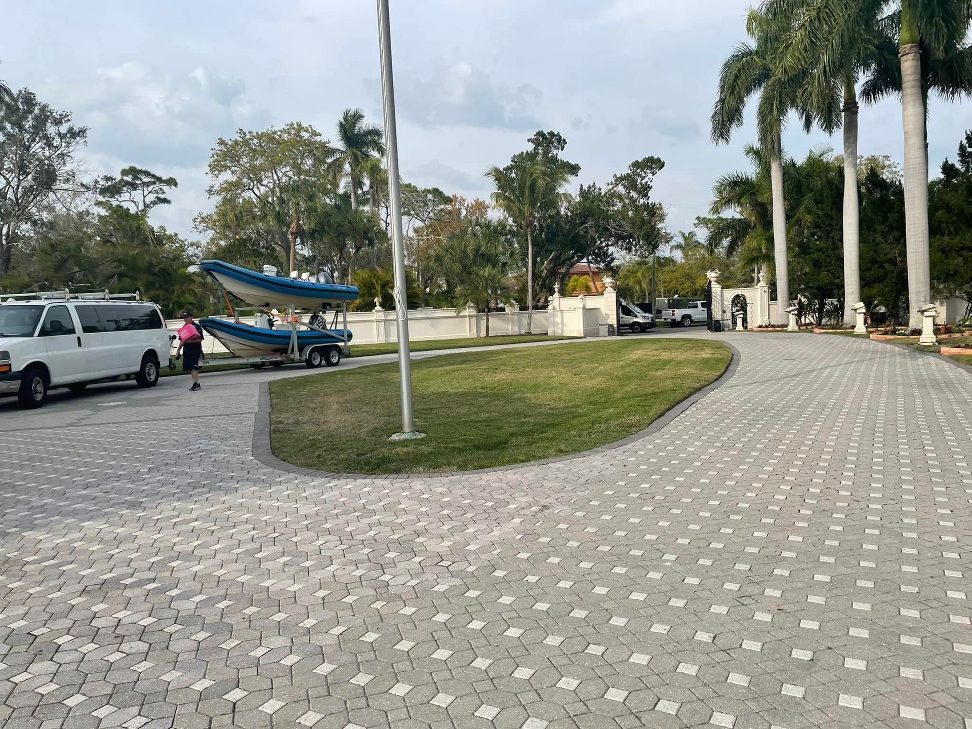 A boat on a trailer, van, and brick driveway at a gated property with palm trees.