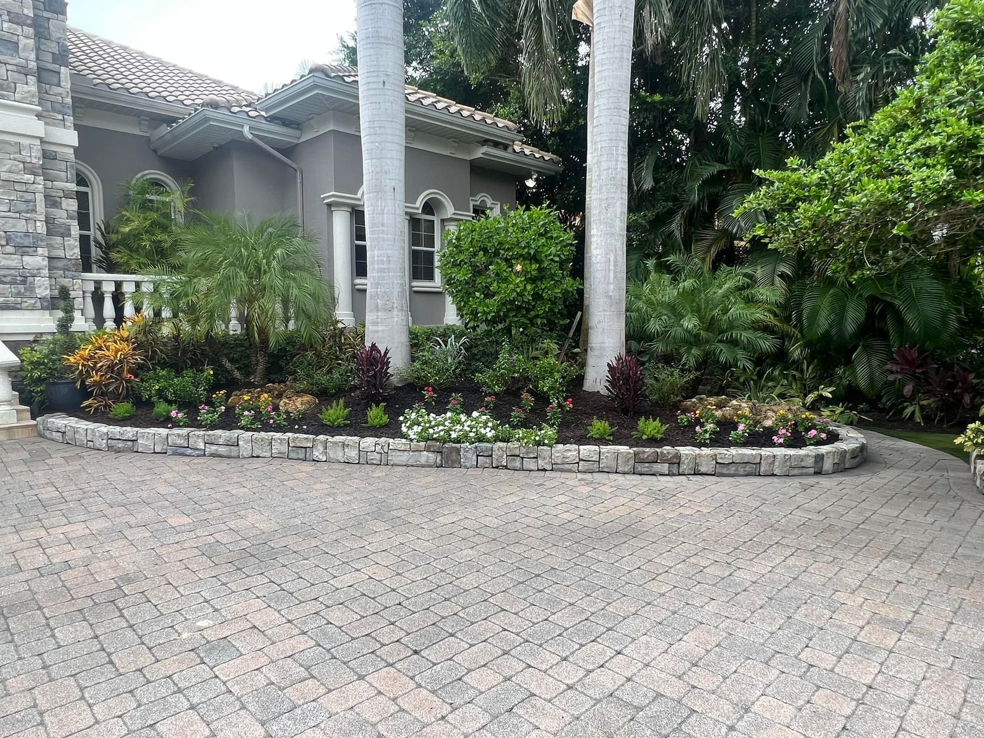 A landscaped flowerbed with a stone border in front of a house. Palm trees and other greenery are featured.