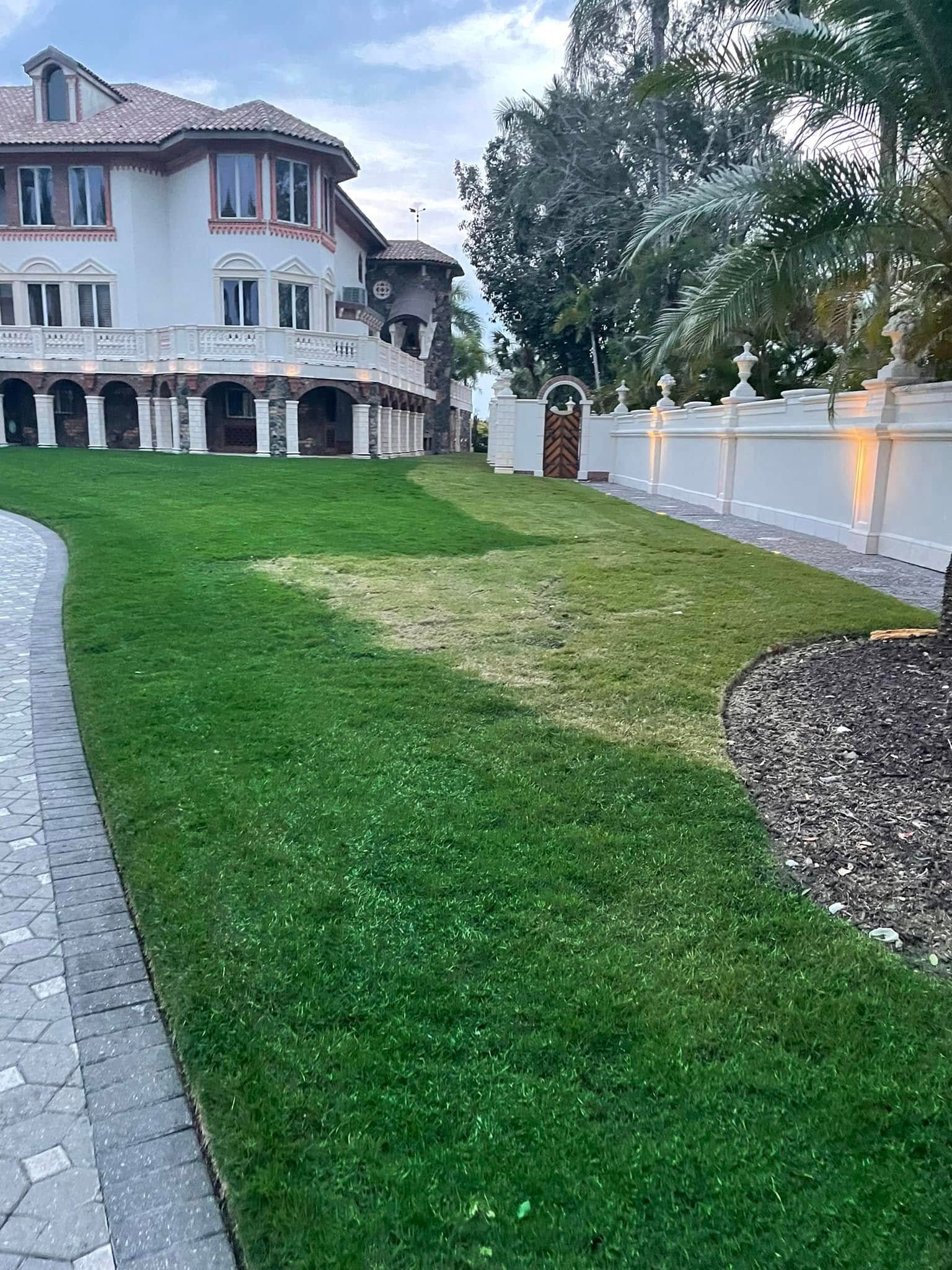 Lush green lawn in front of a large, multi-story white house with columns and a decorative wall.