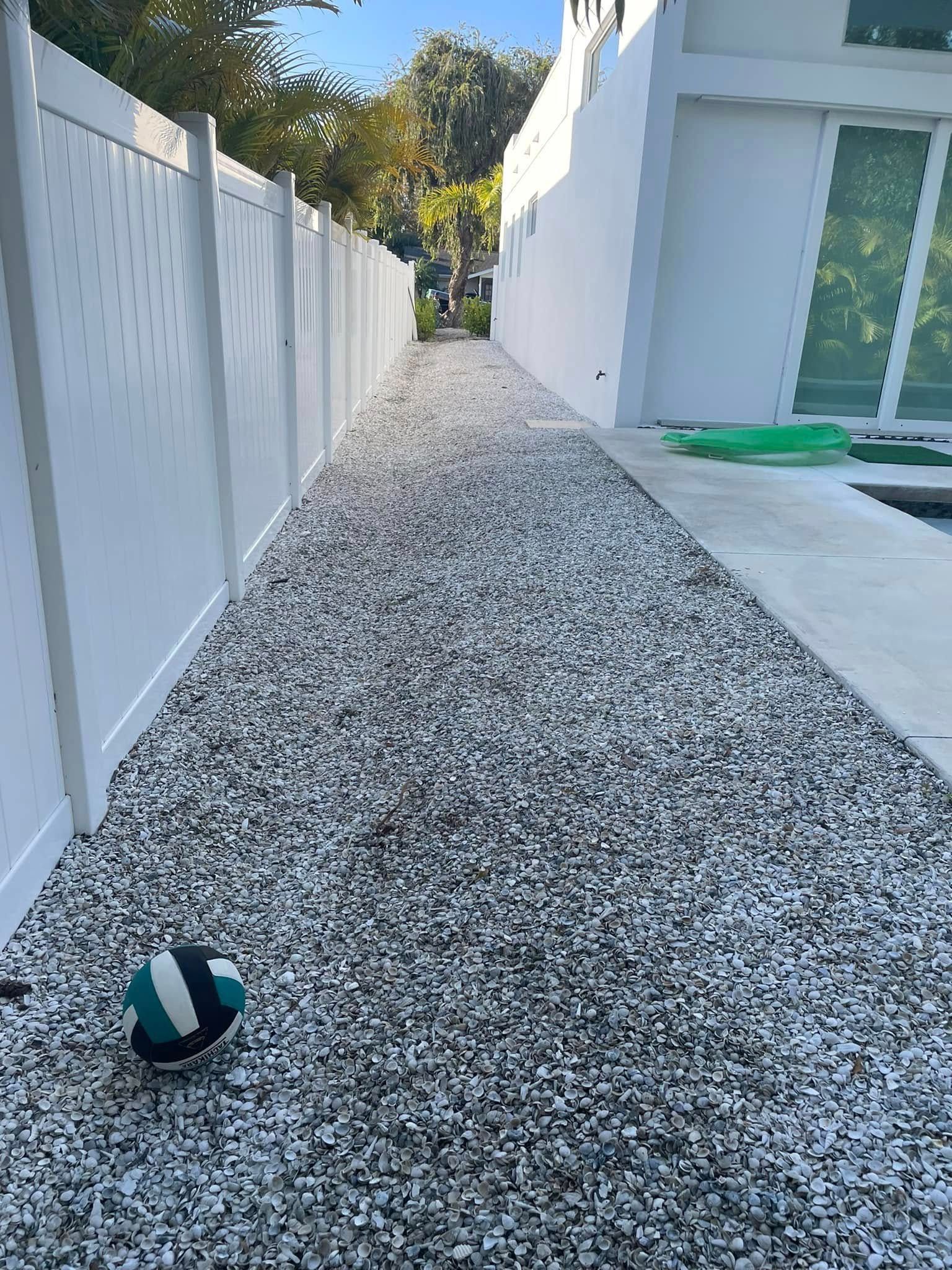 Narrow gravel pathway between a white fence and a white building, with a ball at the bottom.