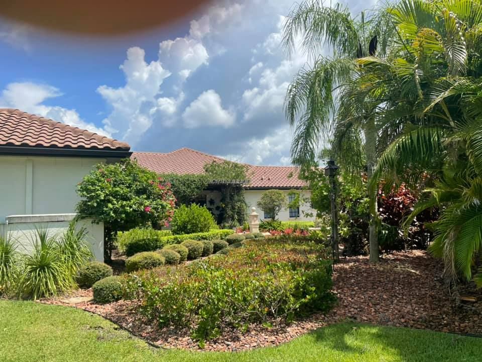 House with a tiled roof, surrounded by landscaped gardens with lush greenery, under a cloudy blue sky.