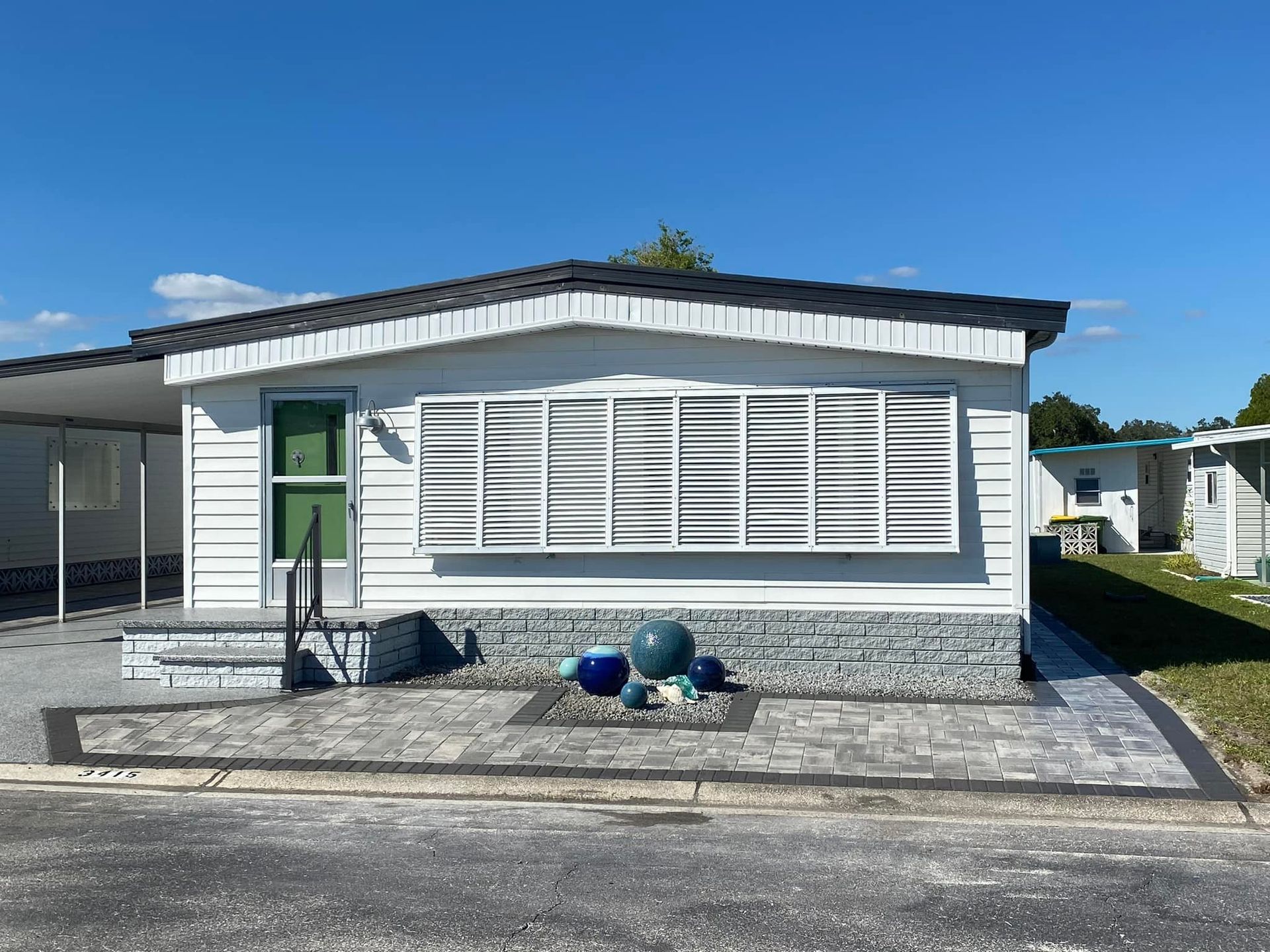 White mobile home with gray accents and decorative blue spheres on front patio, sunny day.