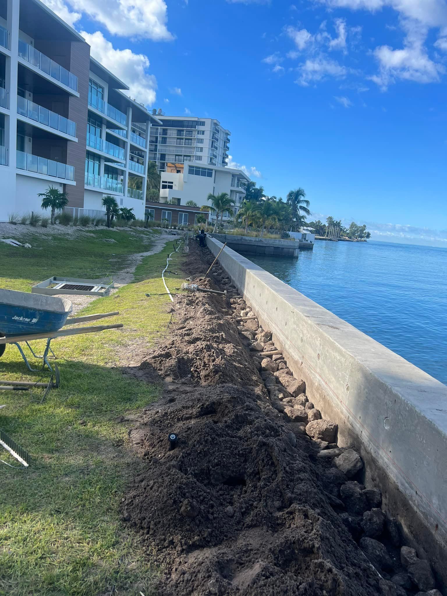 Dirt alongside a concrete waterfront wall with buildings and water in the background. Bright blue sky.