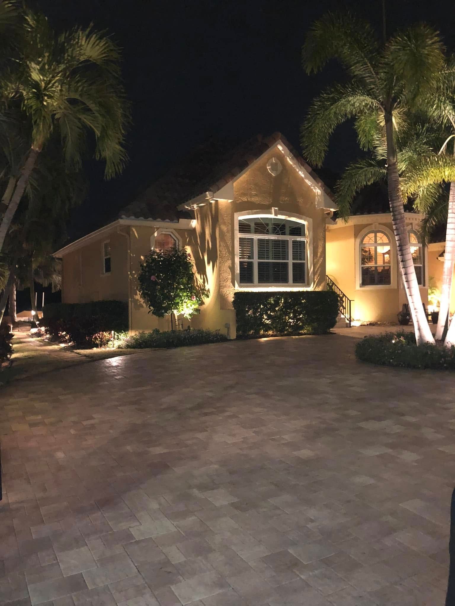 Night view of a house with illuminated landscaping and palm trees. Driveway in the foreground.