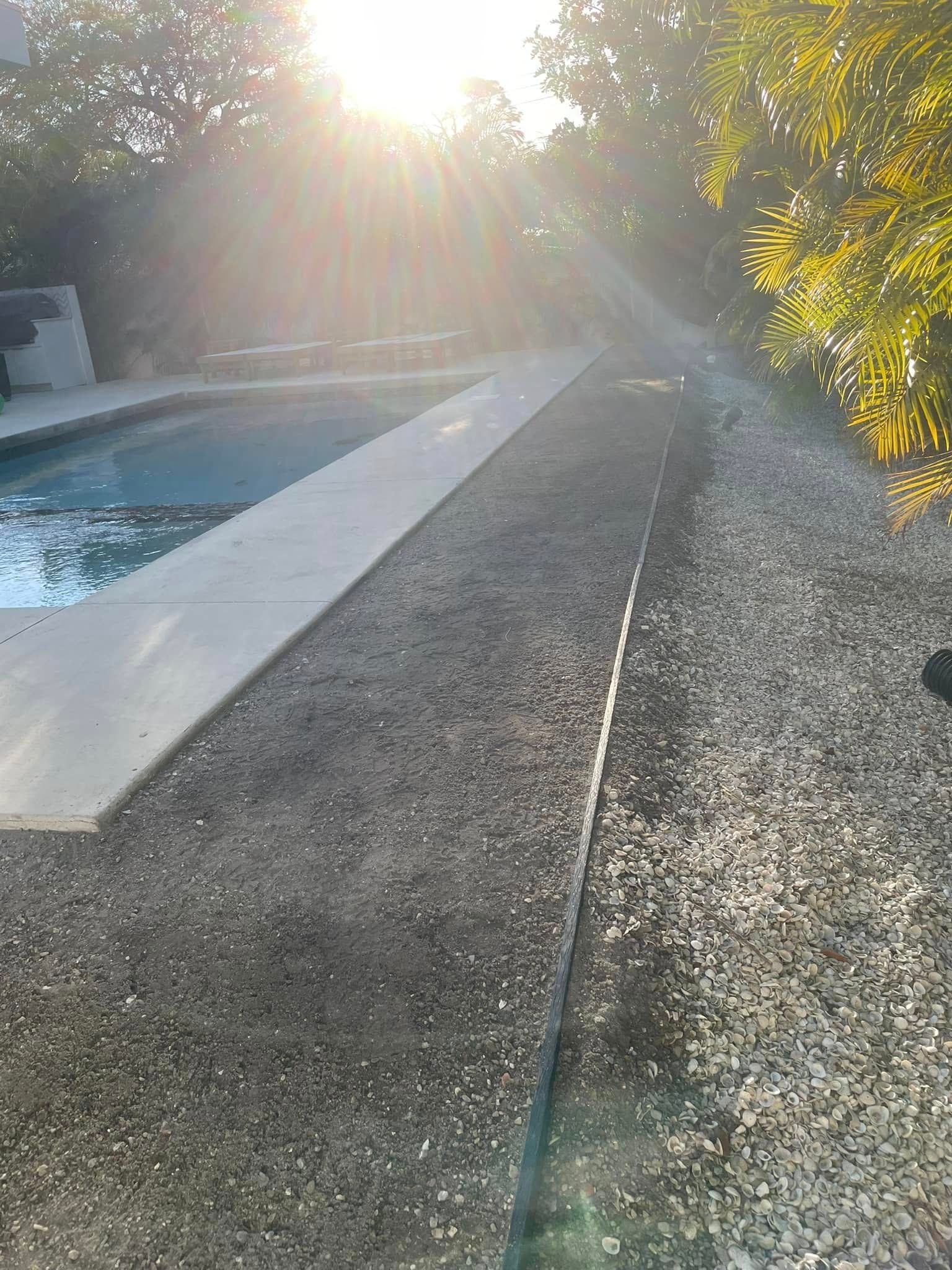 Concrete path beside a pool with small gravel and trees, sun shining.