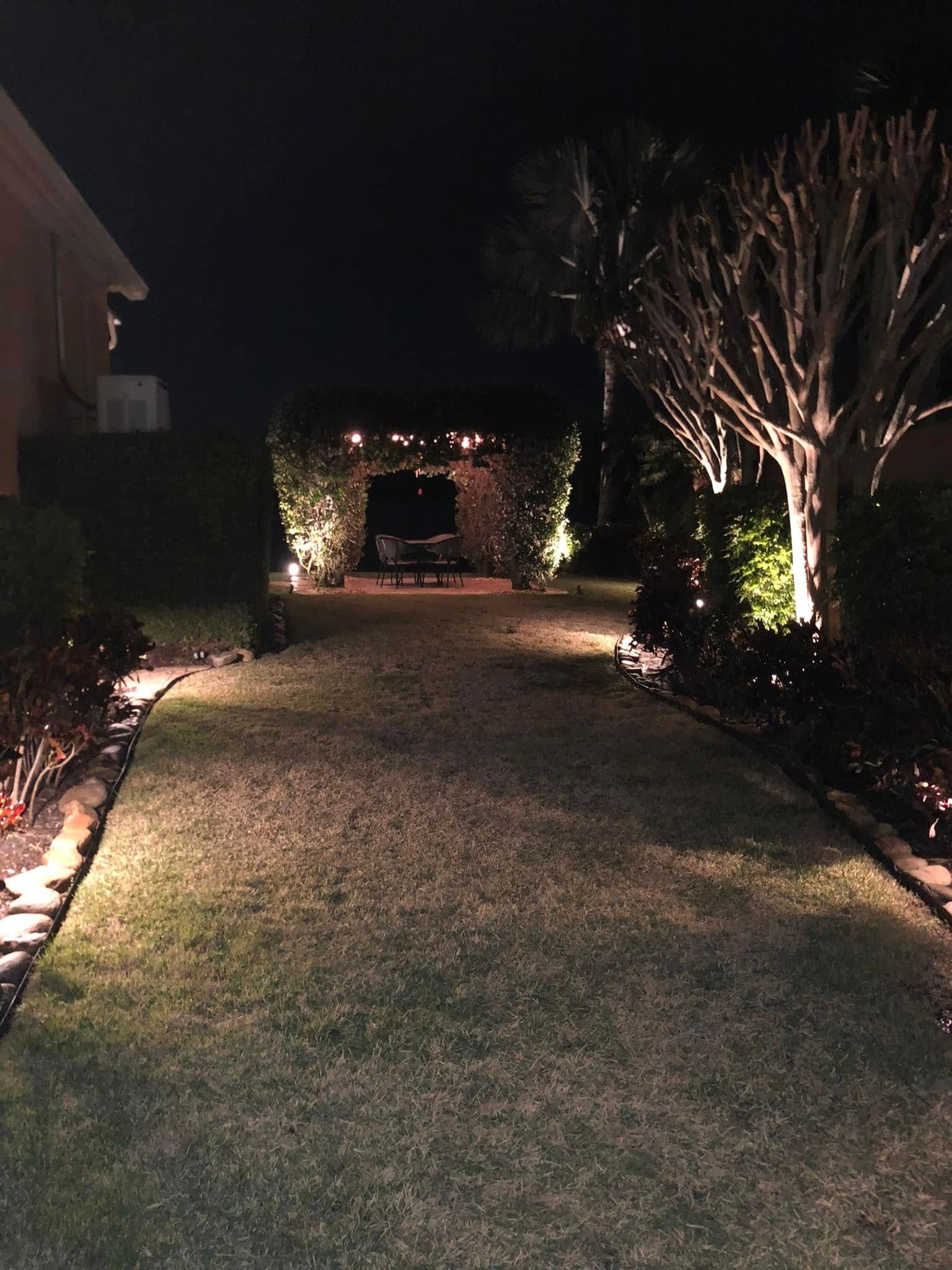 Nighttime view of a well-lit garden path. Warm lights illuminate the grass, hedges, and trees, creating depth.