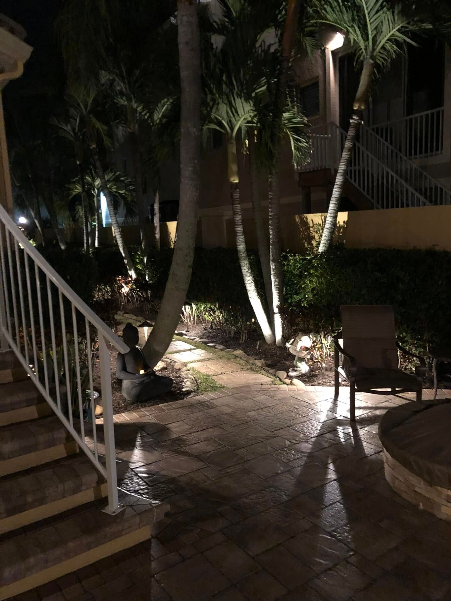 Nighttime outdoor scene with lit palm trees, stone pathway, and a staircase with a white railing.