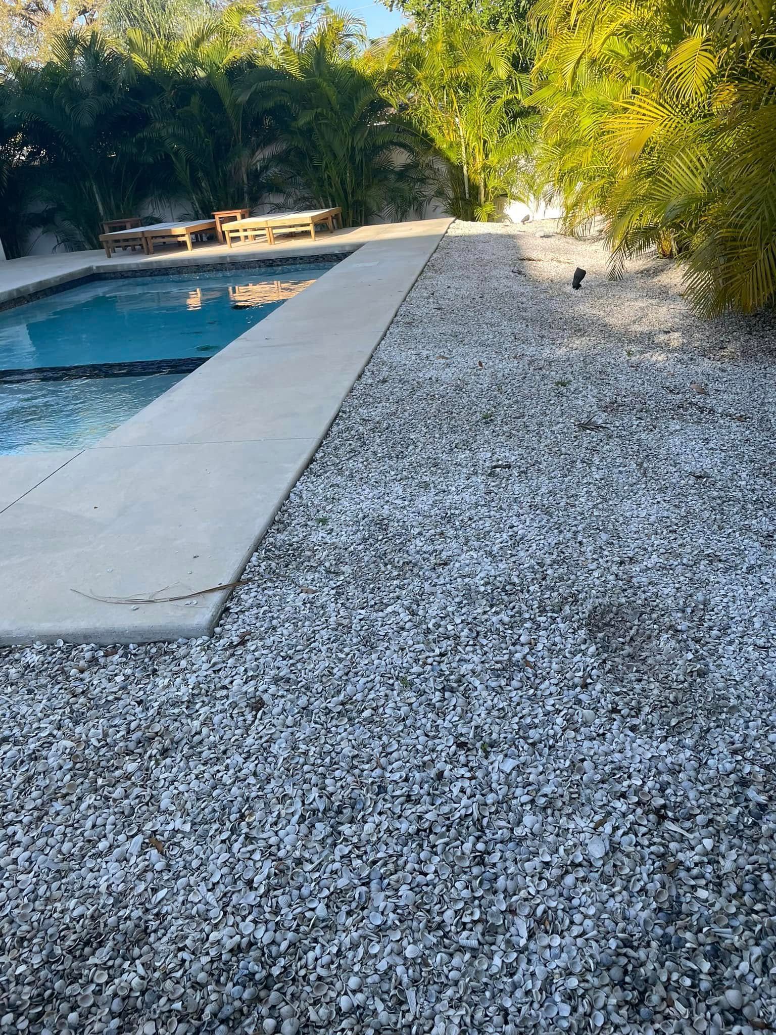 Poolside area with concrete walkway, white gravel, and greenery.