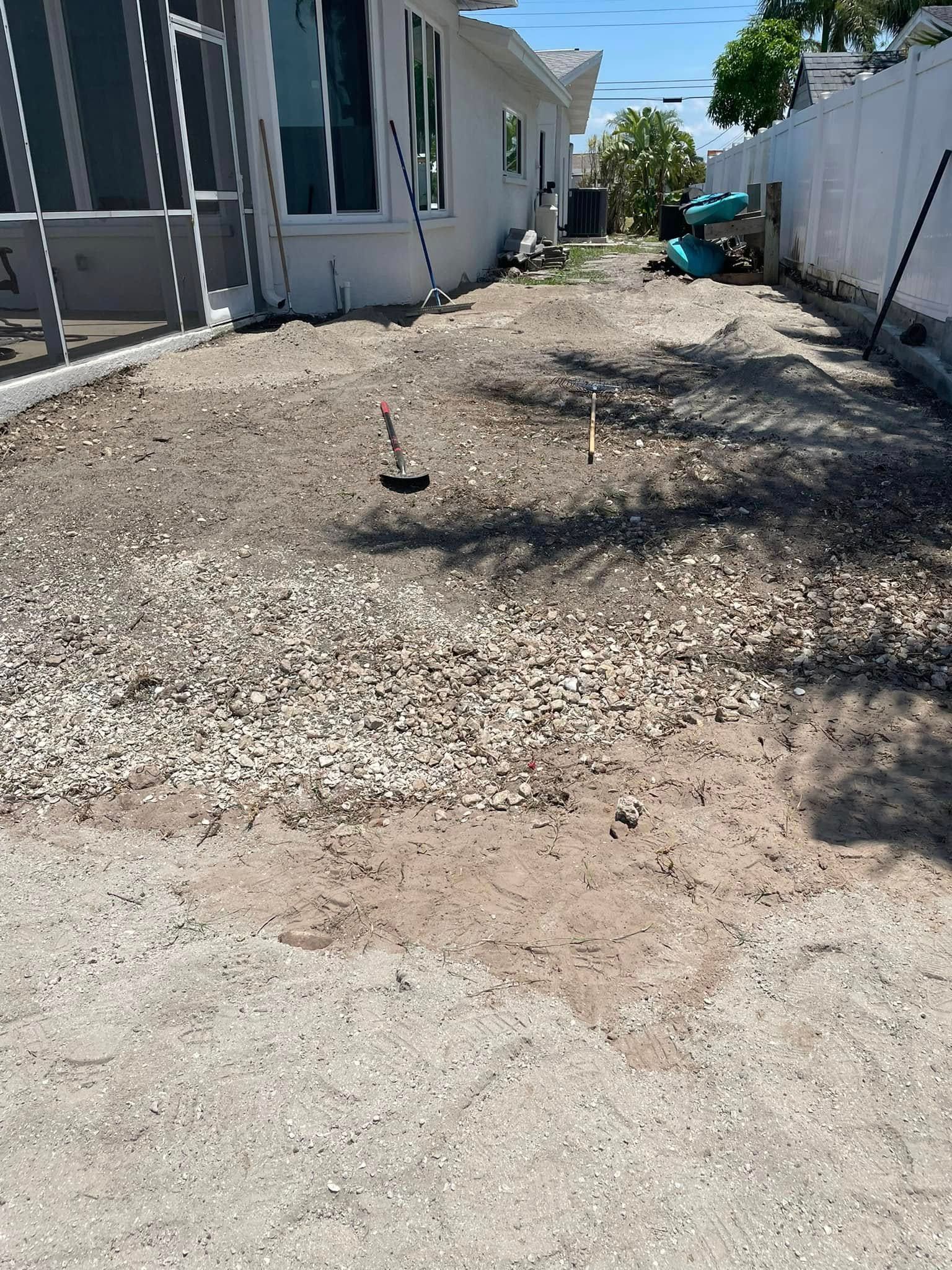 Backyard gravel and dirt area next to a white house and fence; a broom and stakes are on the ground.