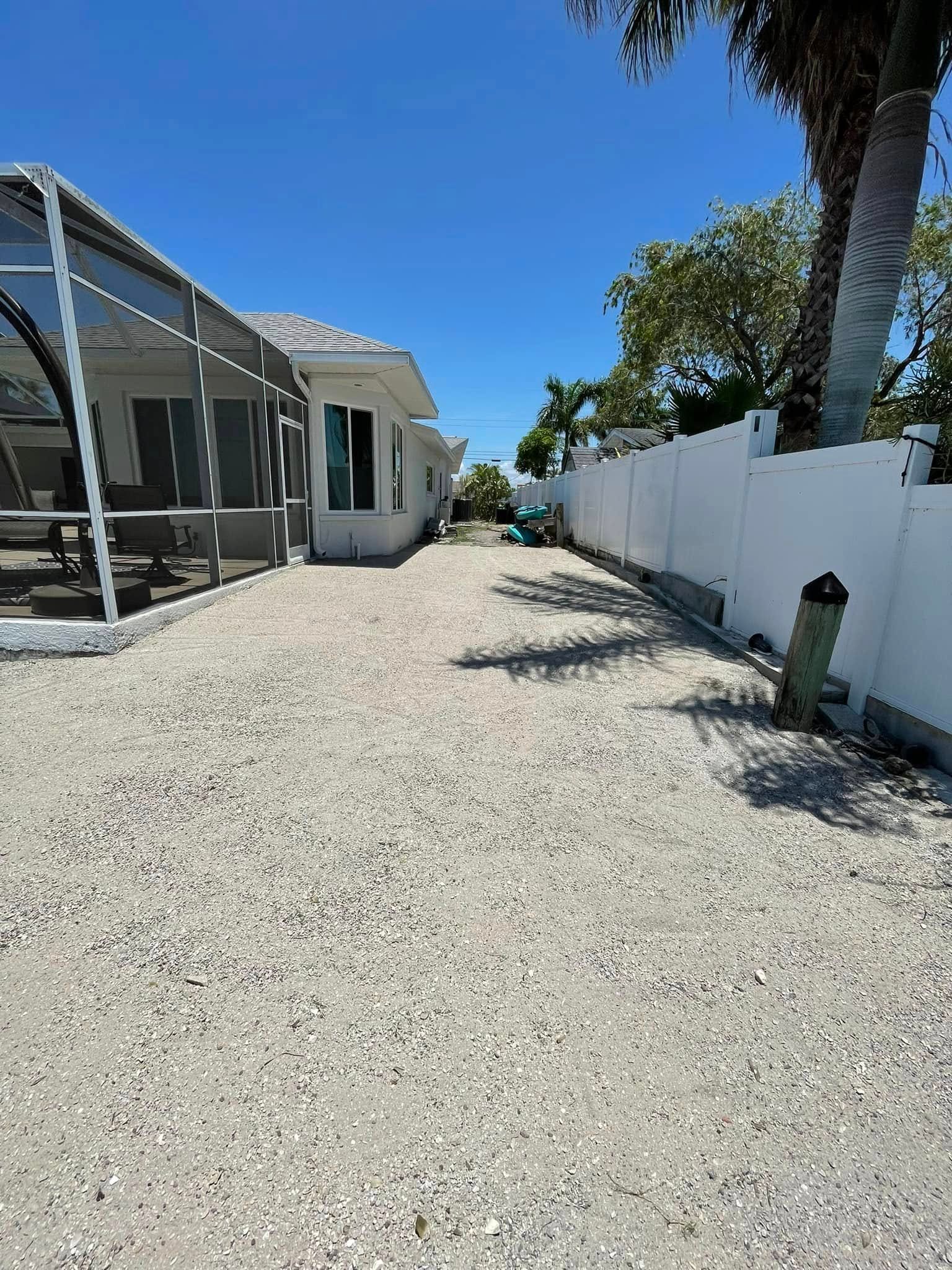 A shell-covered yard alongside a white fence and house under a blue sky.