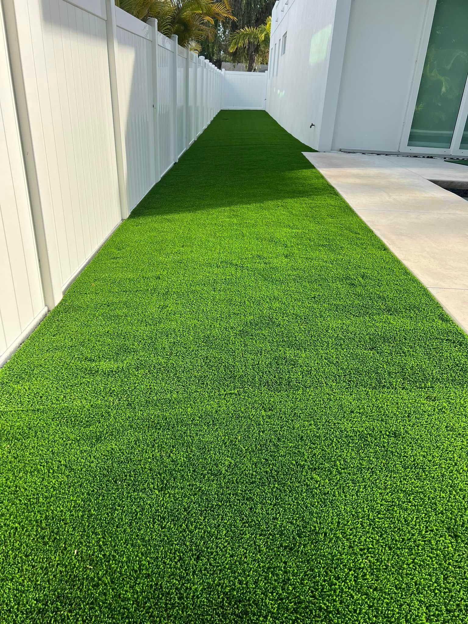 Narrow strip of artificial green turf between a white fence and a white building.