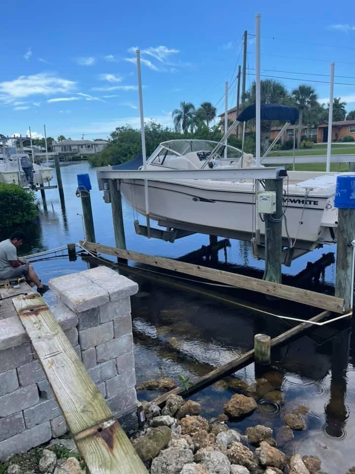 Boat on a lift over water near a dock, blue sky. Person on dock, palm trees in background.