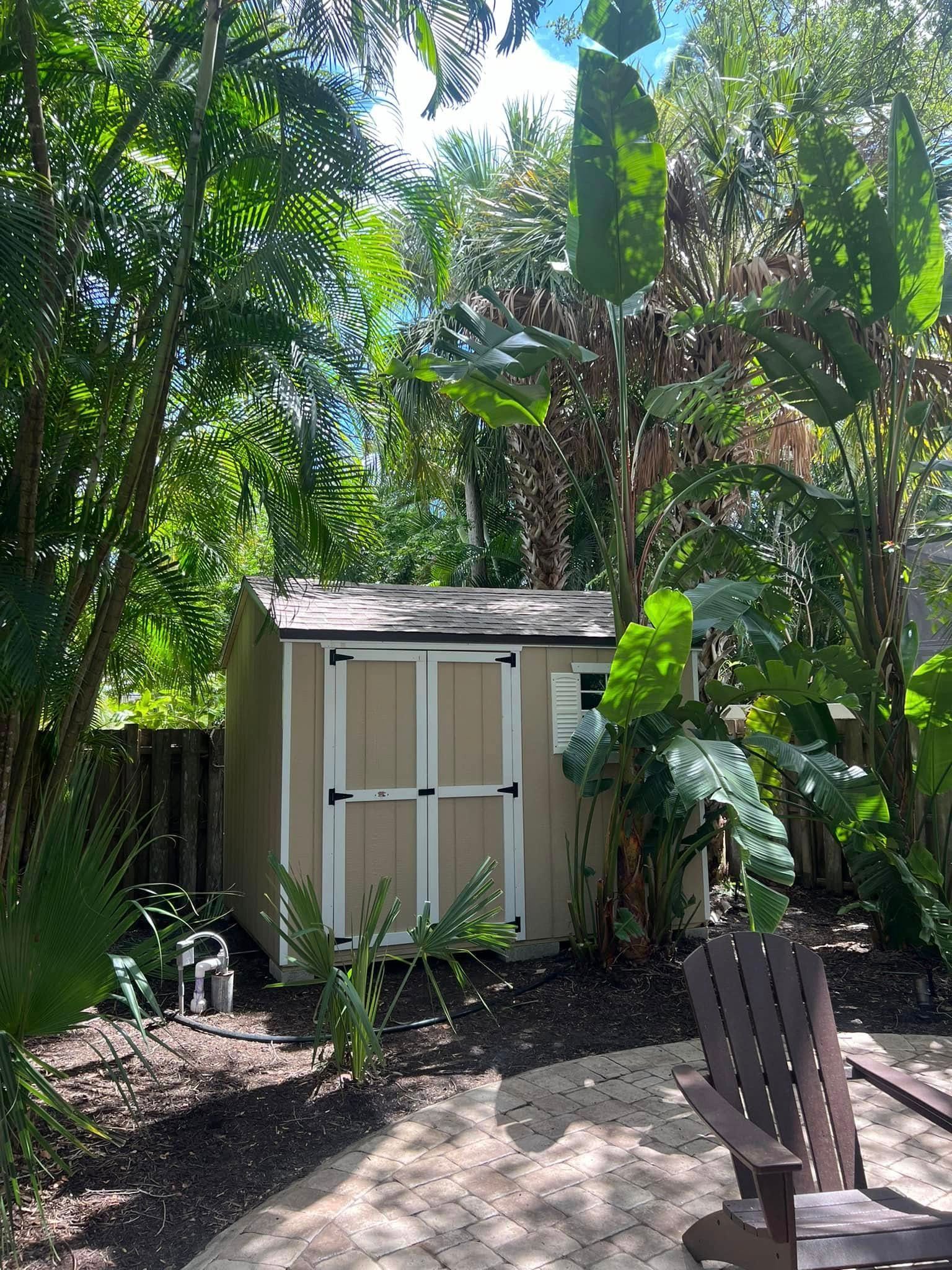 Tan shed surrounded by green trees and foliage in a backyard setting; a patio and chair are visible.