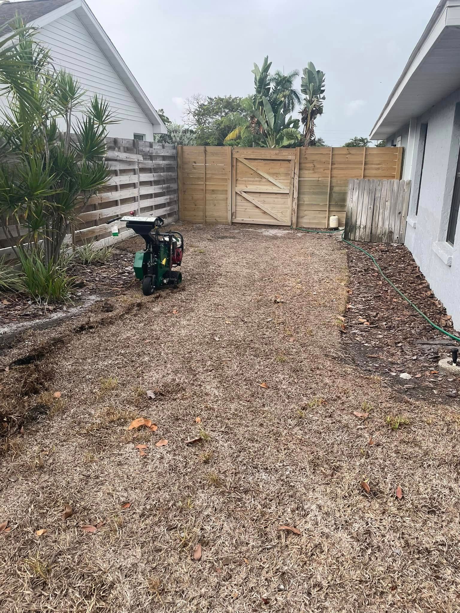 A gravel driveway leads to a wooden fence with a gate. A wood chipper sits near the fence.