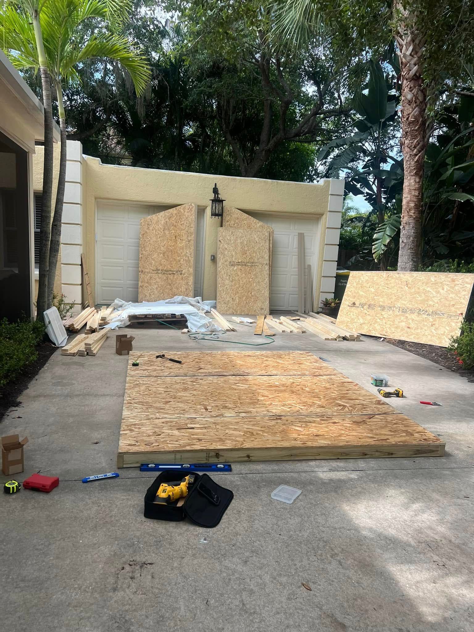Construction materials and tools are scattered on a driveway, near a building with two garage doors.