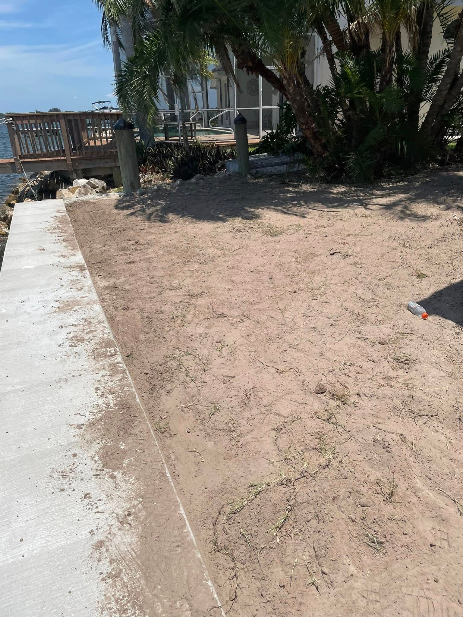 Sandy ground alongside a concrete walkway near waterfront with trees and a deck.