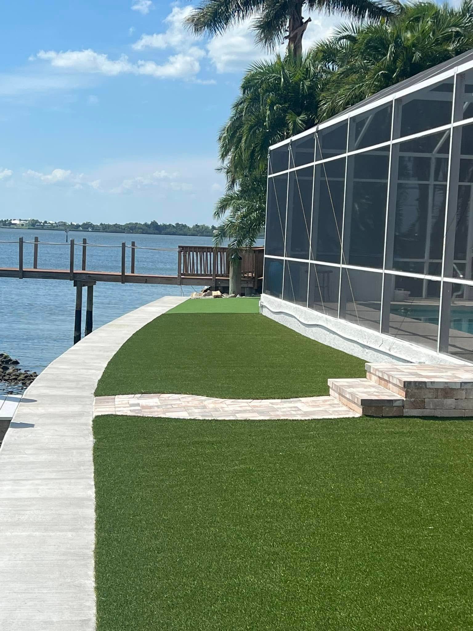 Lush green lawn next to a waterfront with a dock, screened-in pool enclosure, and a bright blue sky.