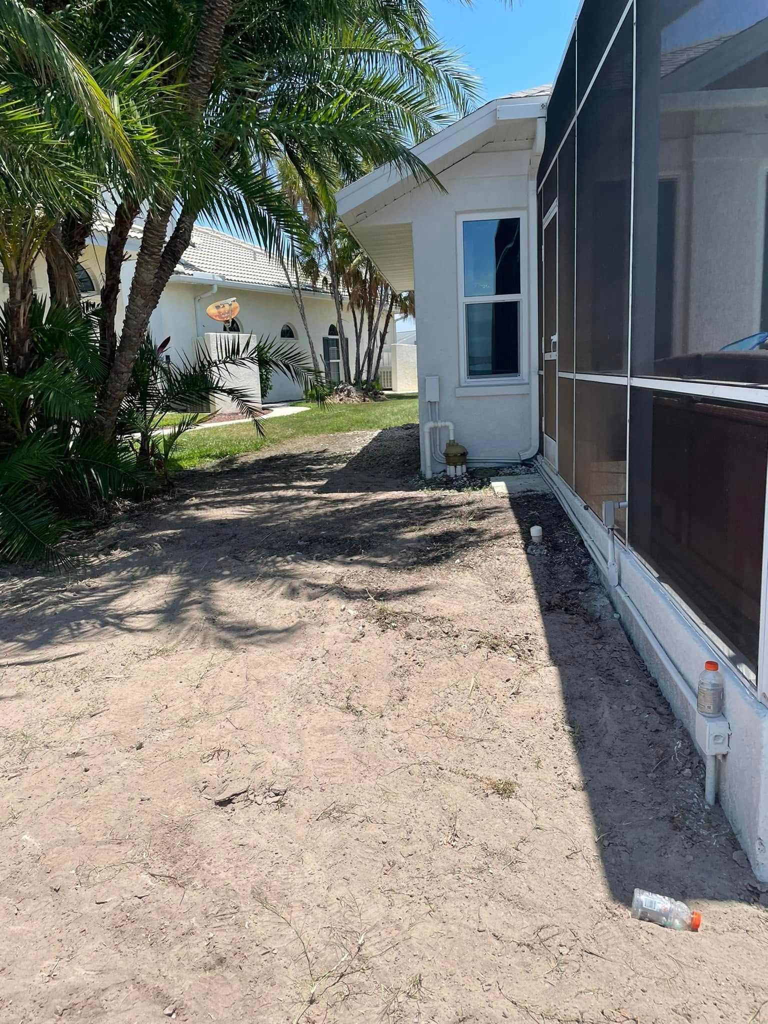 Sandy yard alongside a white house and screened porch, with vegetation and blue sky.