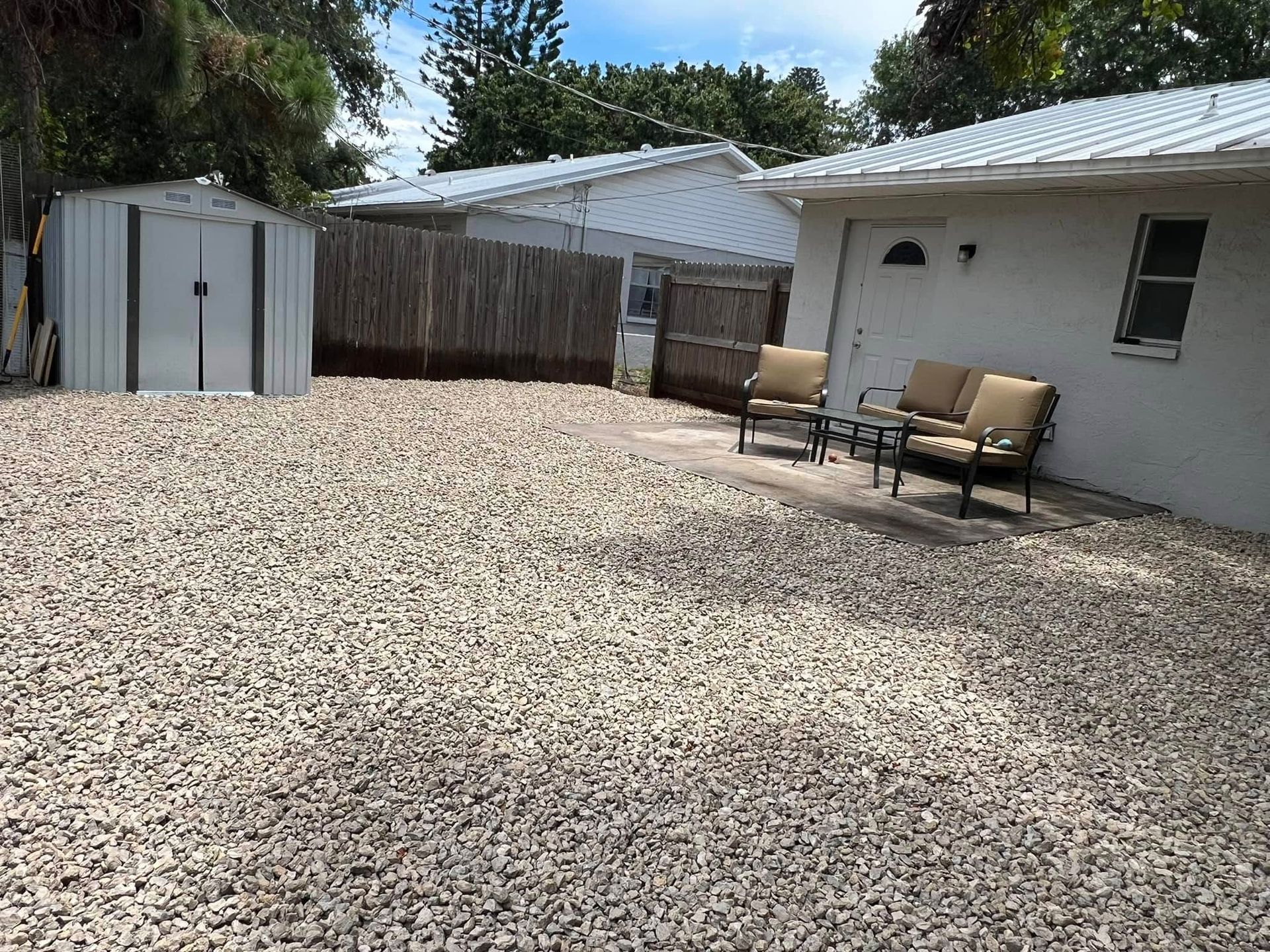 Gravel backyard with shed, fence, and seating. White buildings, blue sky.