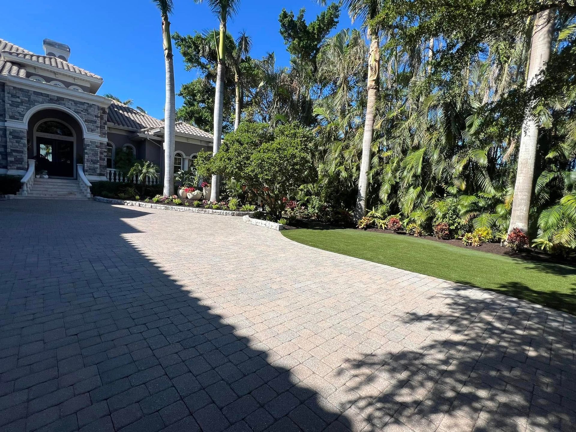 Driveway leading to a stone house with palm trees and landscaping on a sunny day.